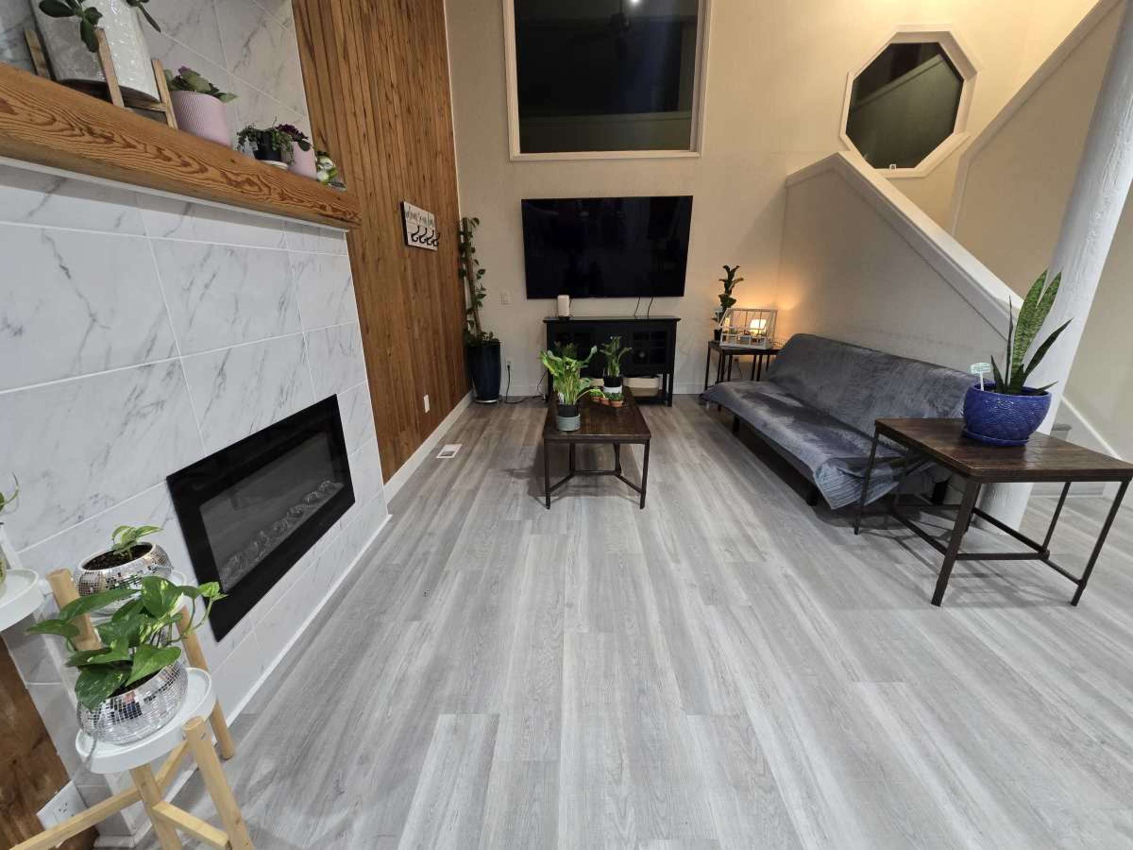 The image shows a spacious living room featuring a fireplace, a grey sofa, a coffee table, and various indoor plants.