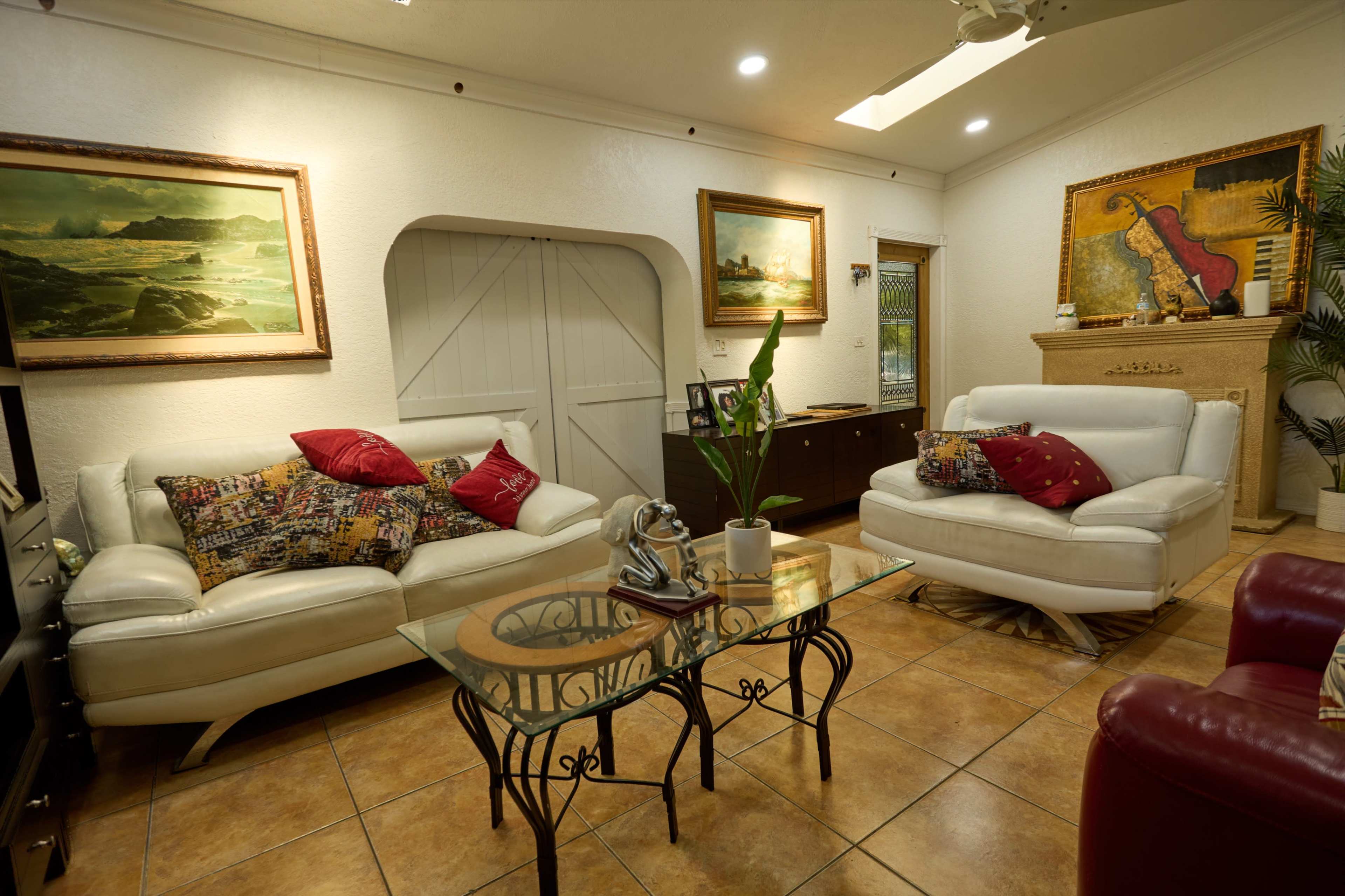 The image shows a cozy living room with white leather sofas, a glass coffee table, and paintings on the walls.