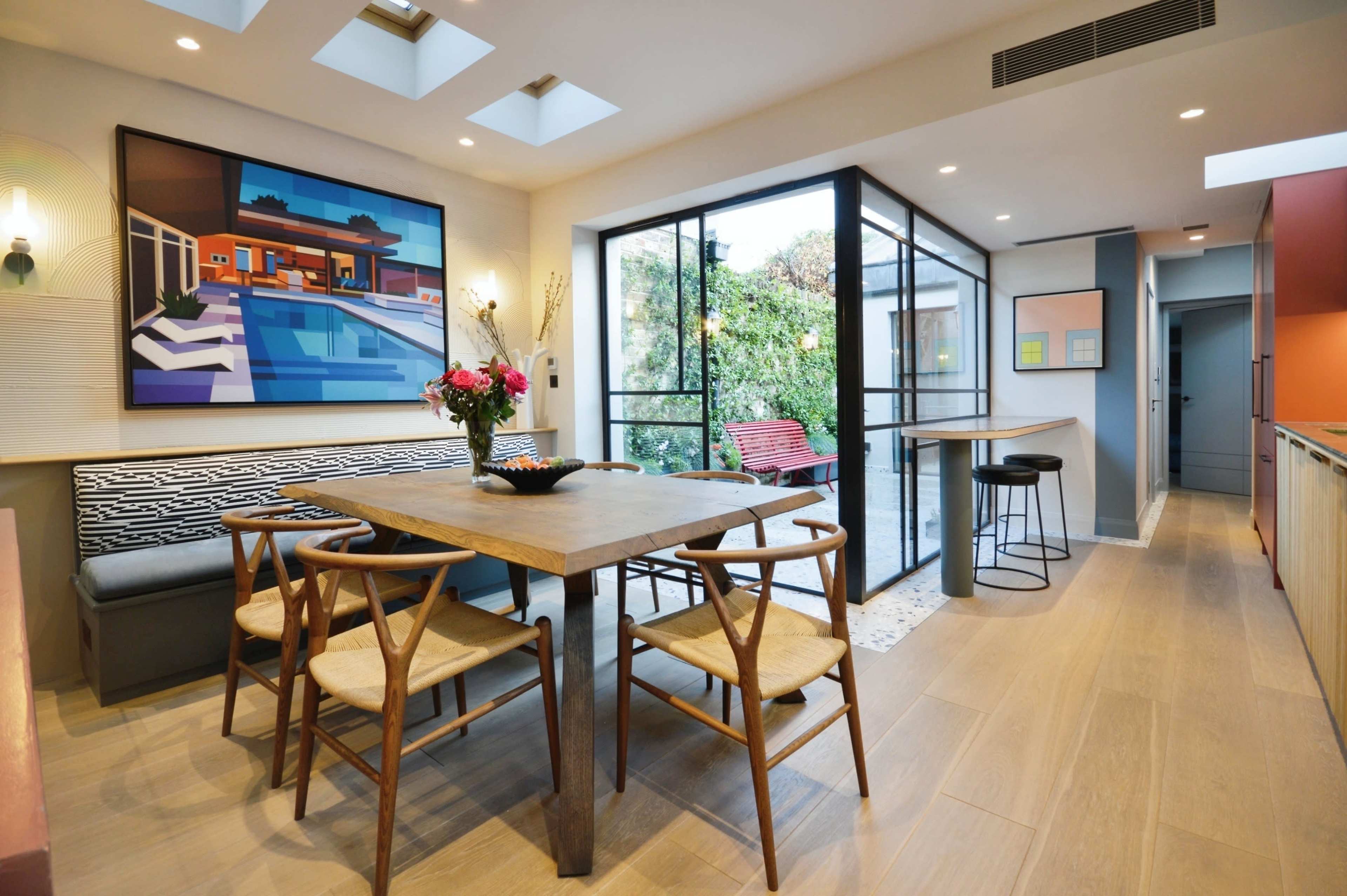 The image shows a modern dining area with a wooden table surrounded by chairs, large glass doors leading to an outdoor space, and a colorful artwork on the wall.