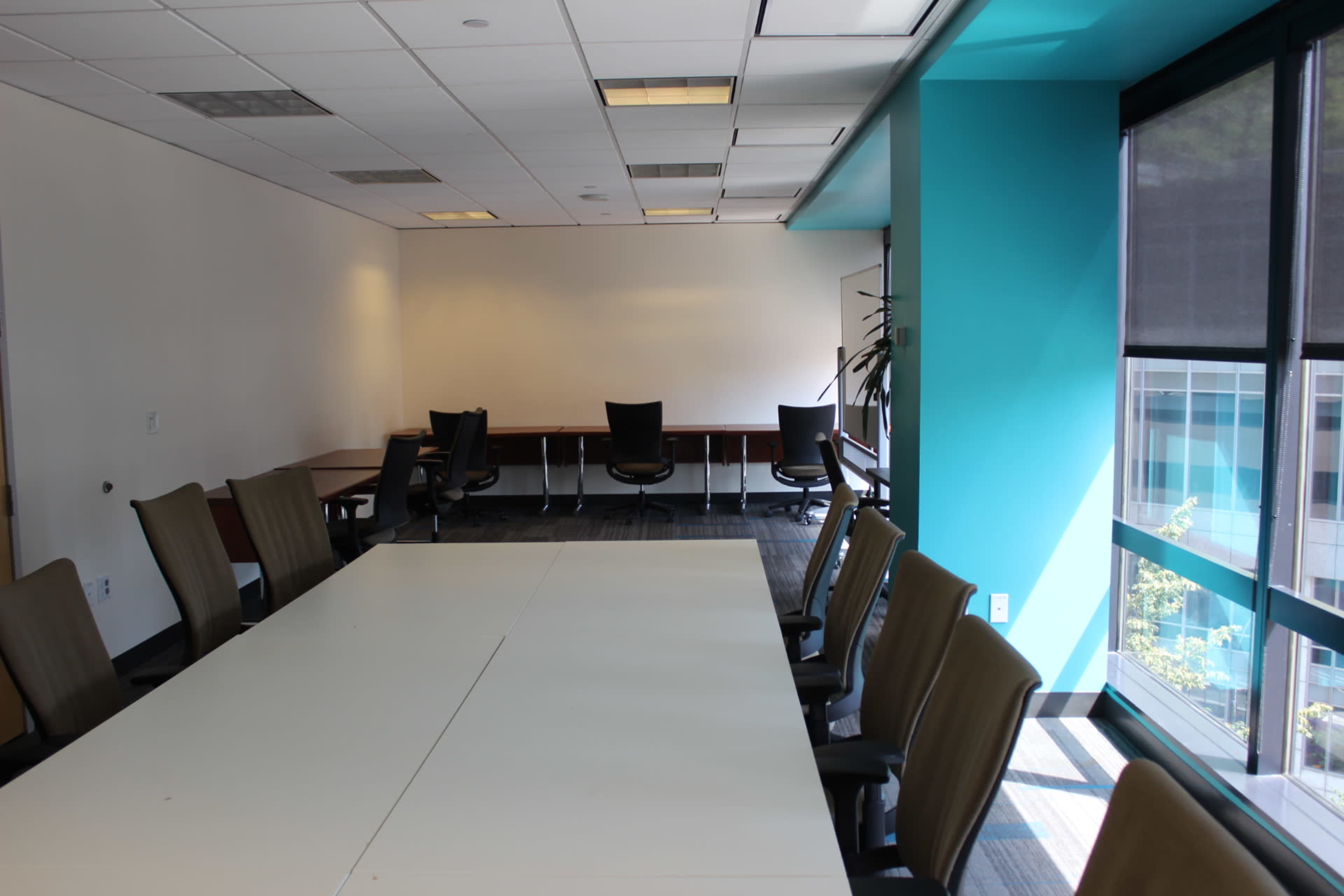 A modern conference room features a long table with several chairs, a large whiteboard, and windows offering a view of the outside.
