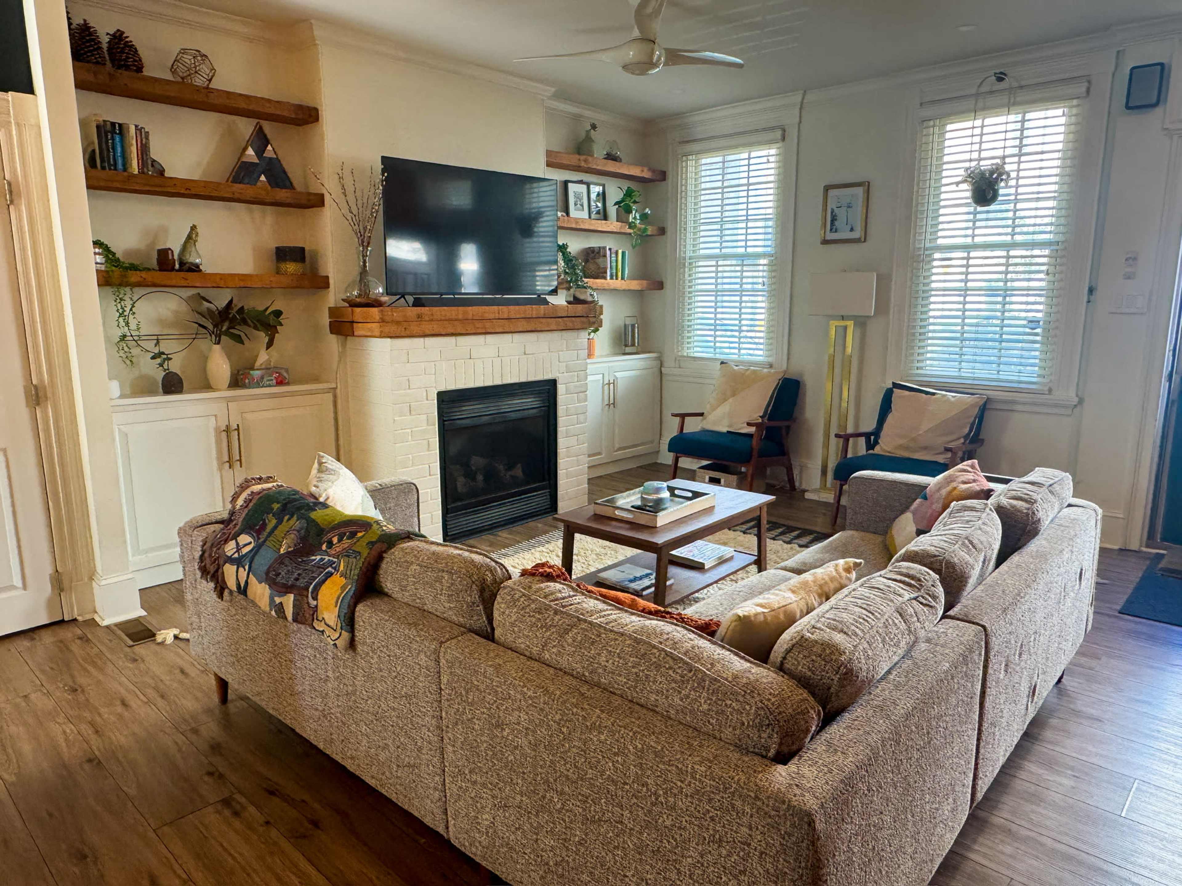 A cozy living room features a beige sectional sofa, a coffee table, a fireplace, shelves with plants and books, and two blue chairs.