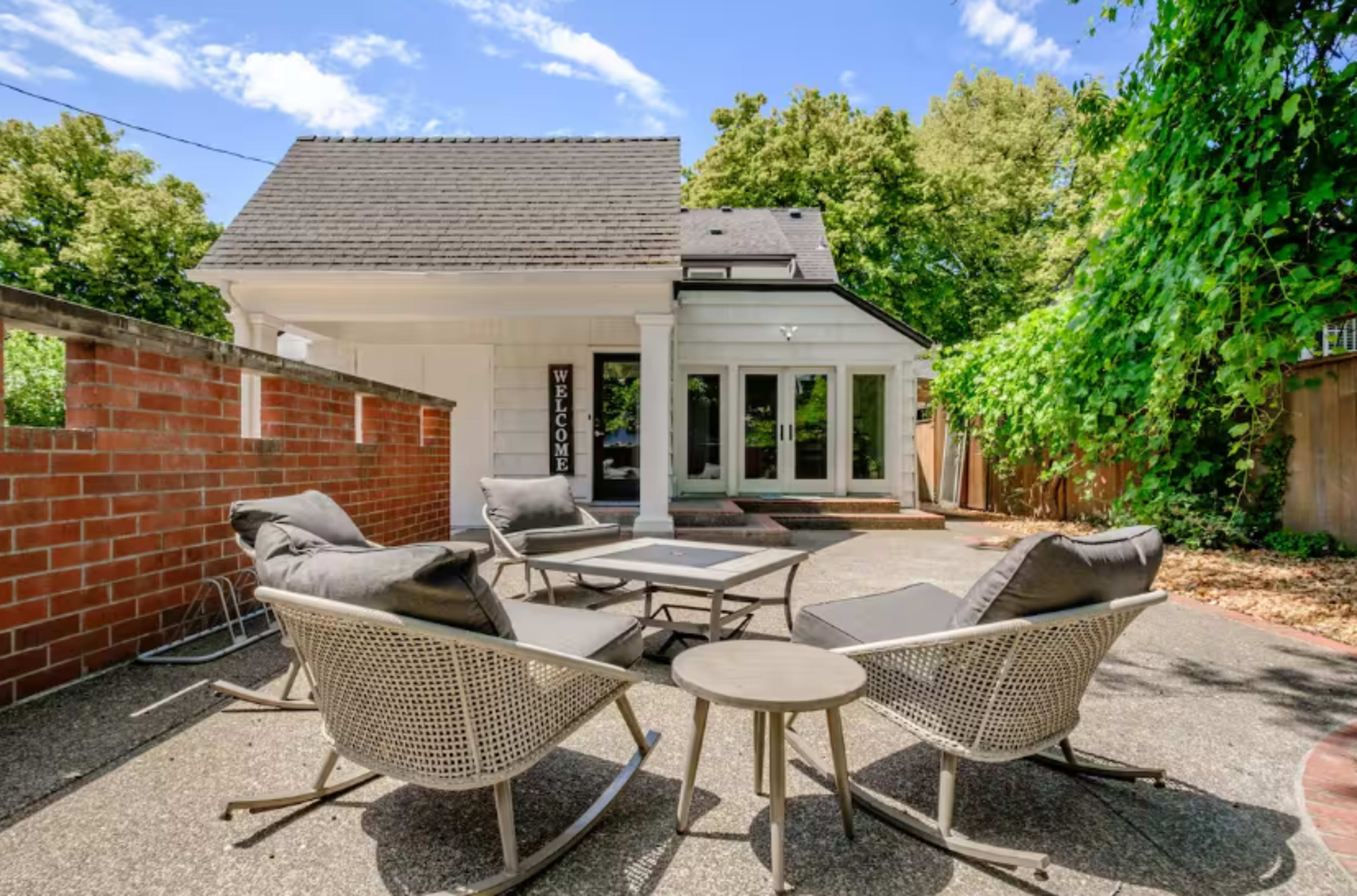 A patio area features modern seating and a table, adjacent to a house with a welcoming entrance and surrounded by greenery.