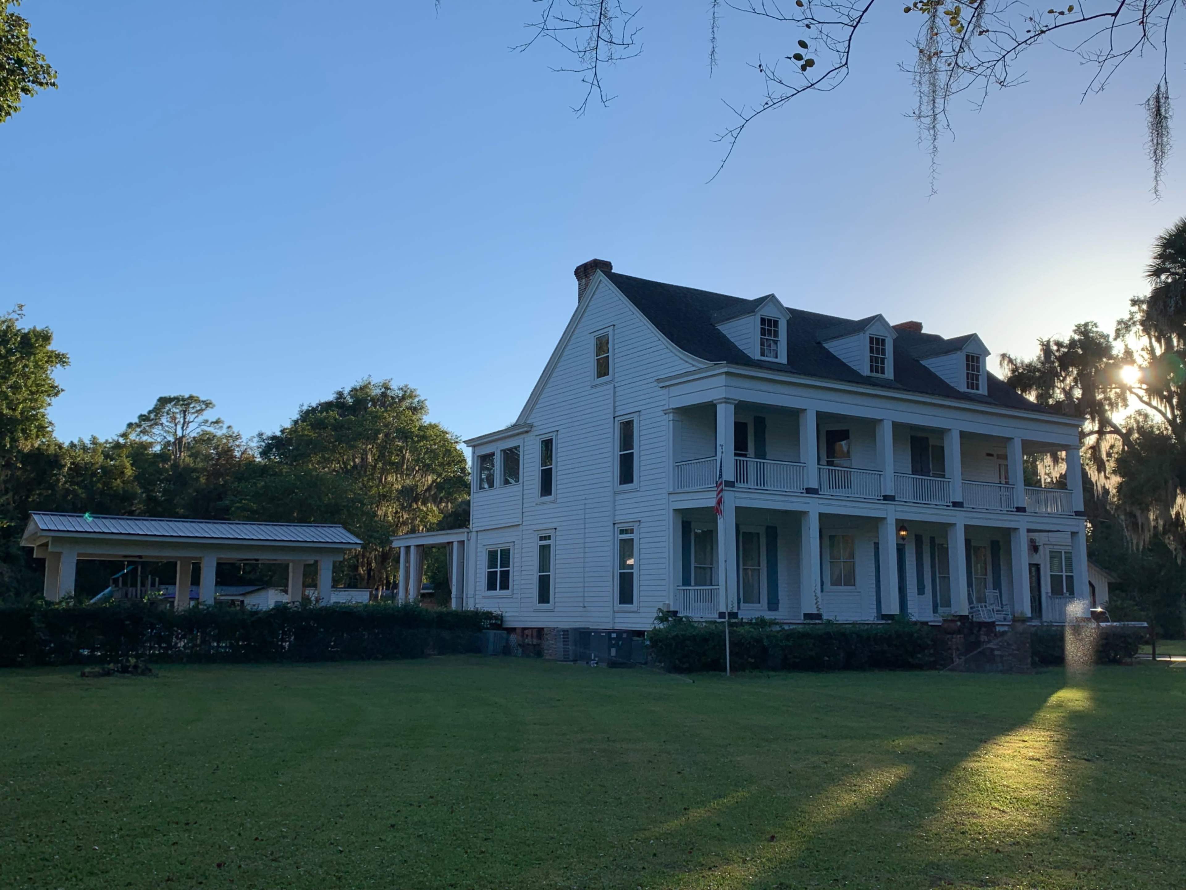 A large, historic two-story house with a front porch and white siding is set on a grassy lawn.