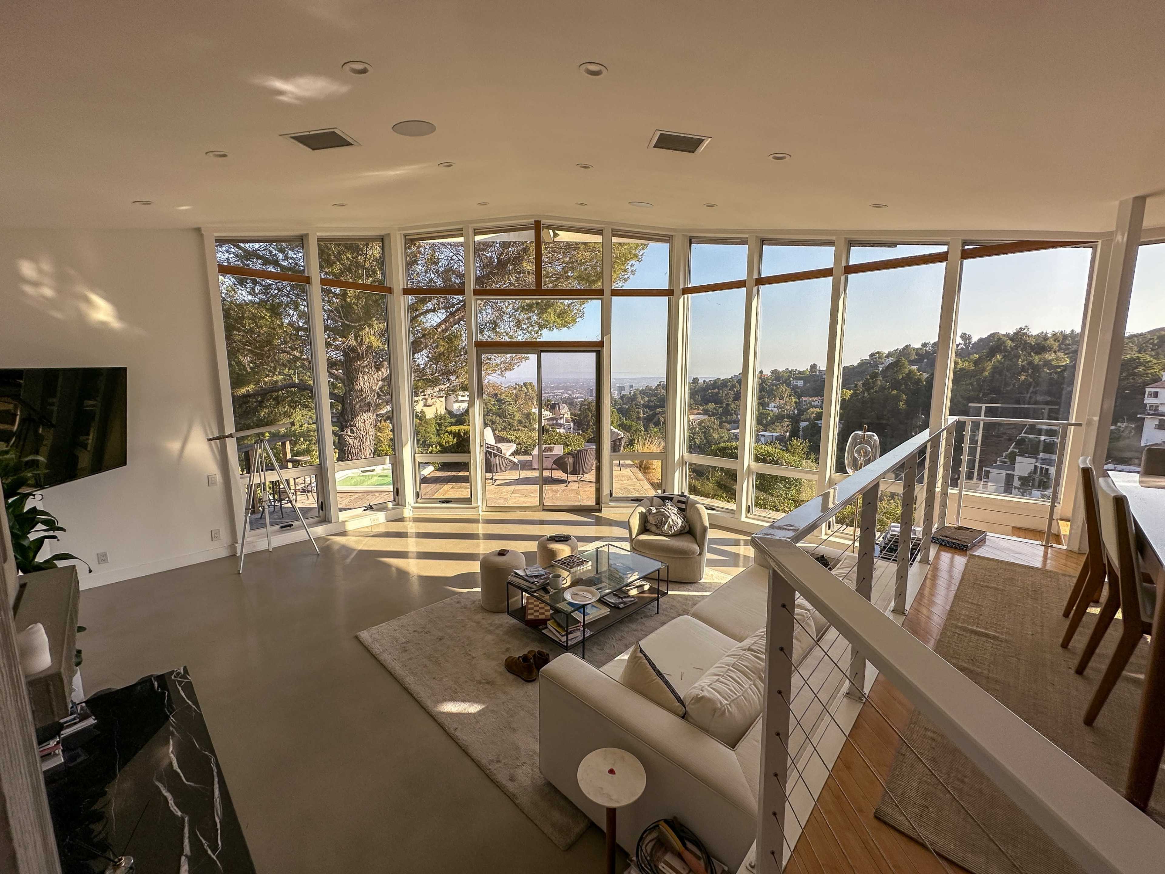 The image shows a modern living room with large glass windows showcasing a view of the hillside and cityscape.