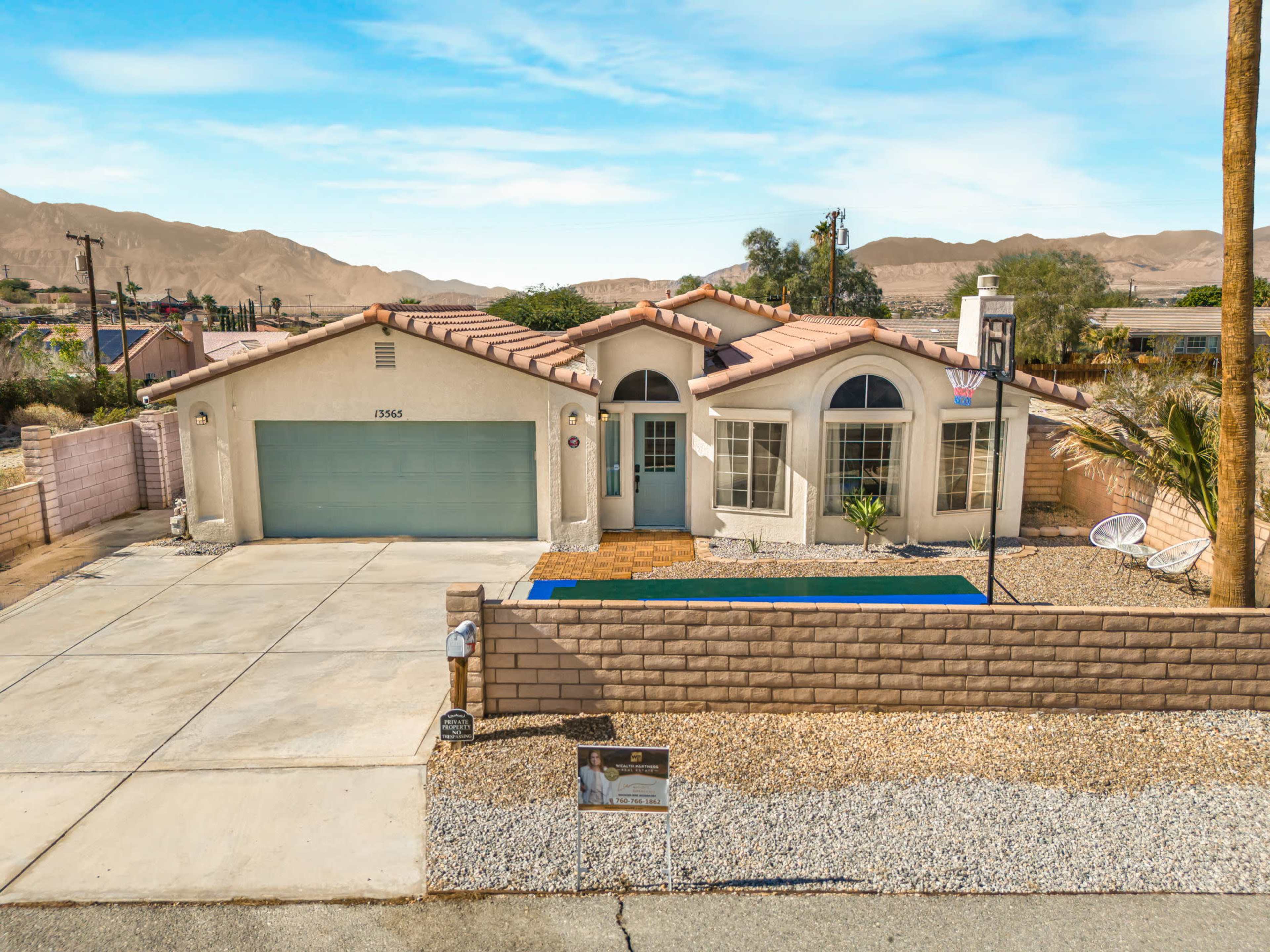 The image shows a single-story house with a curved entryway, a decorative basketball hoop, and a gravel driveway, situated in a arid landscape with mountains in the background.