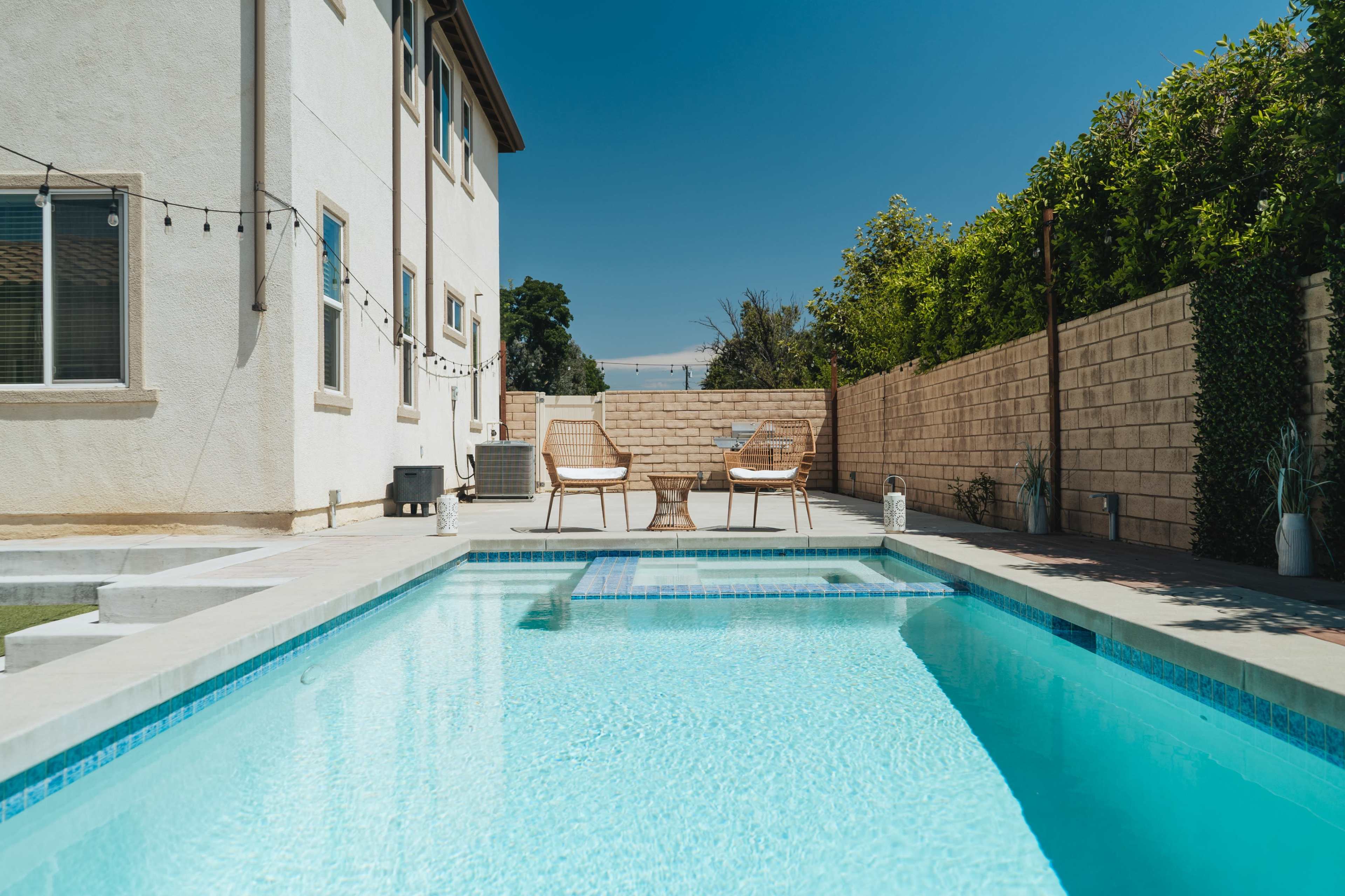 A clear swimming pool is situated beside a modern house with two chairs positioned on the patio and greenery lining the yard.