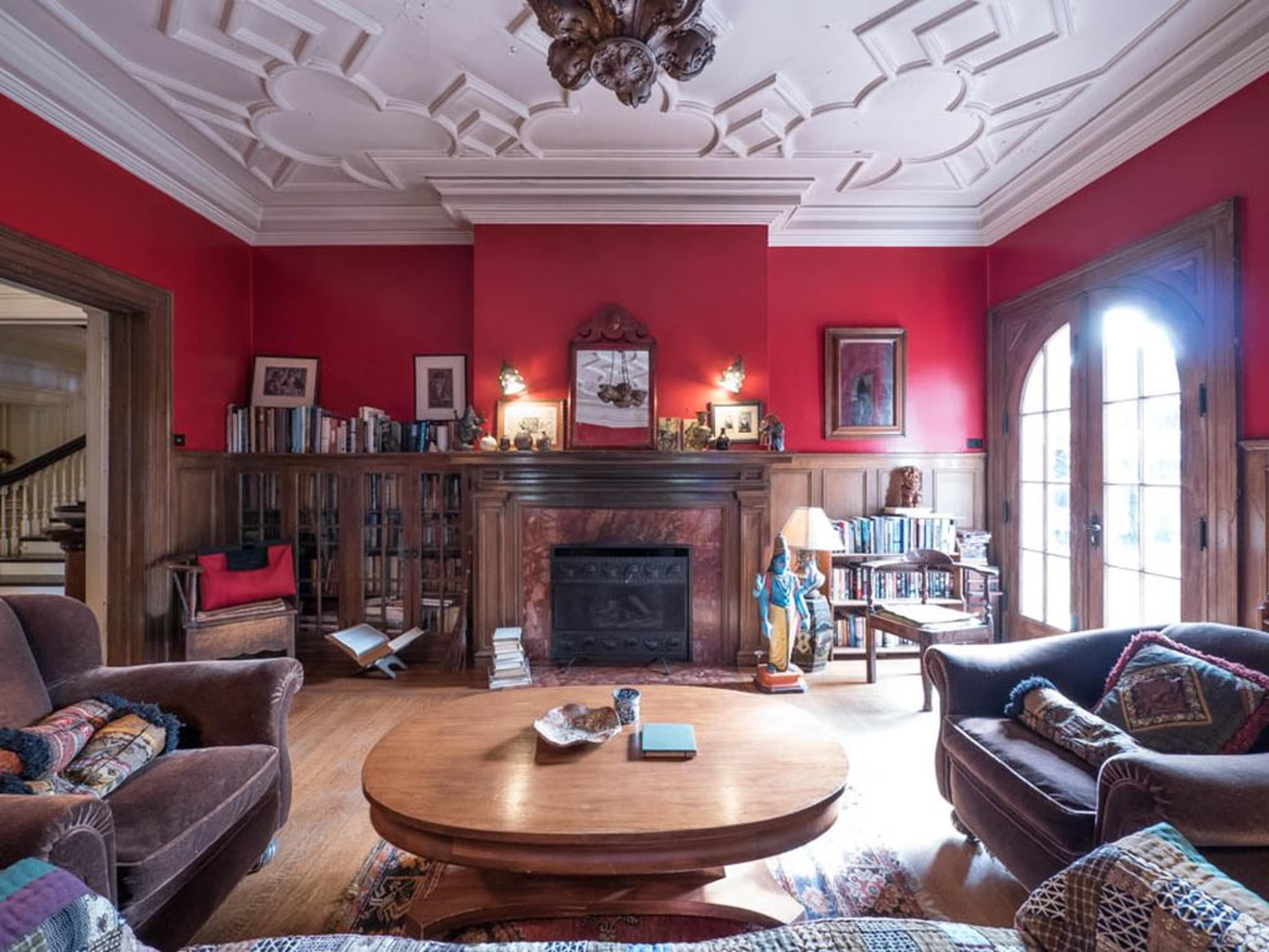 The living room features a red wall, a wooden ceiling with decorative molding, a central round table, and bookshelves filled with books.