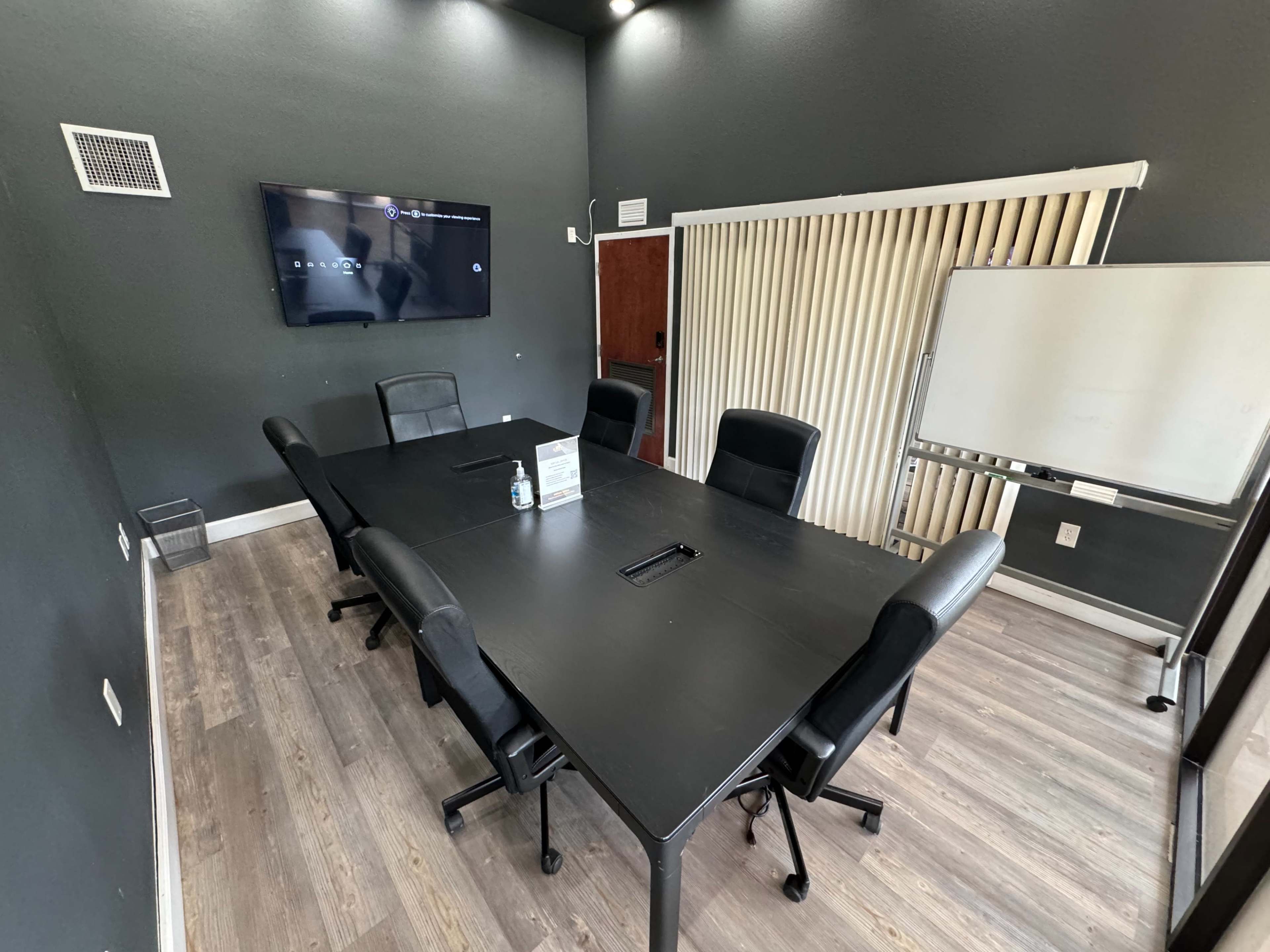A conference room features a large black table surrounded by black chairs, with a whiteboard and a television mounted on the wall.