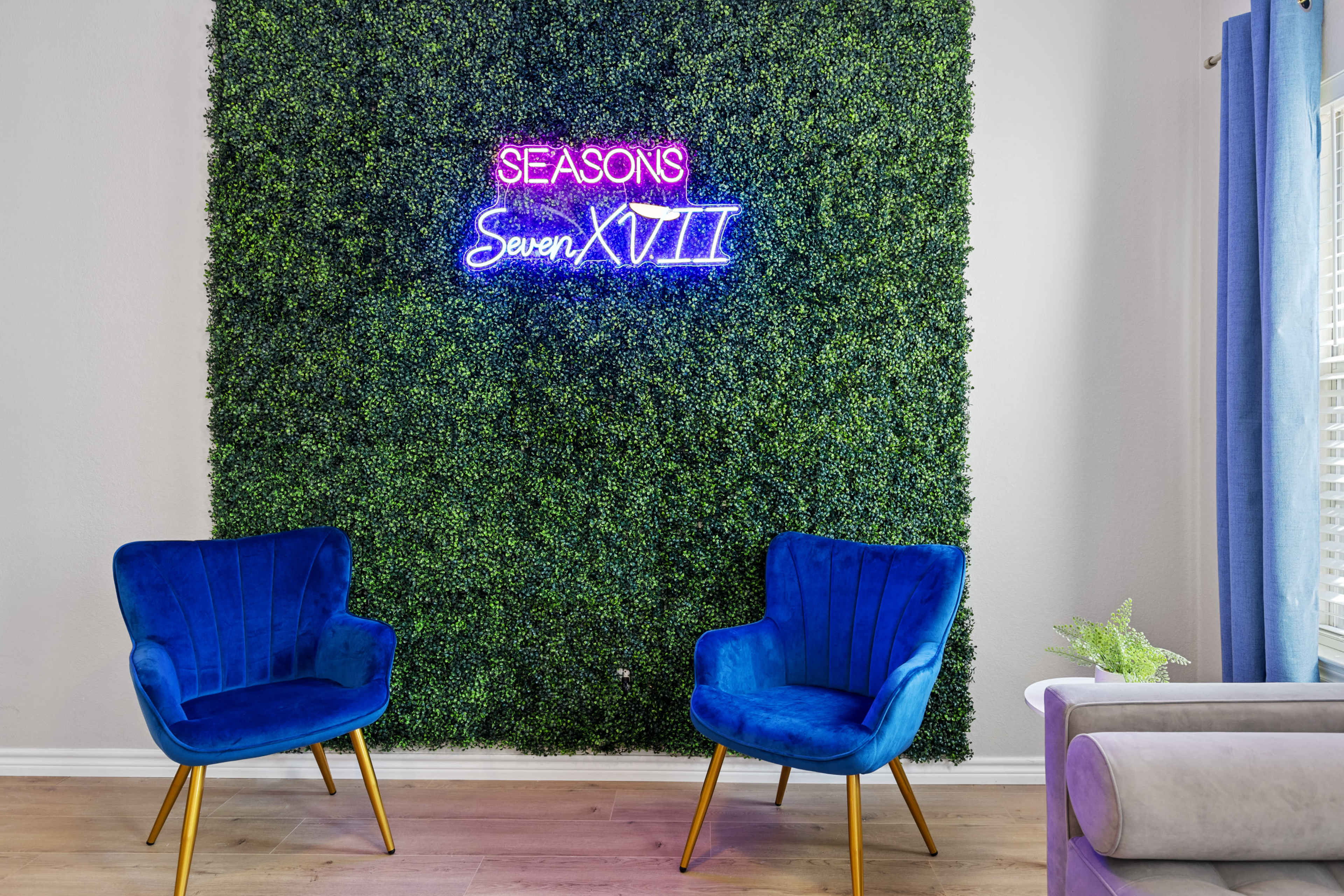 The image features a green wall made of artificial foliage with a neon sign reading "SEASONS Seven XVII" above two blue velvet chairs.