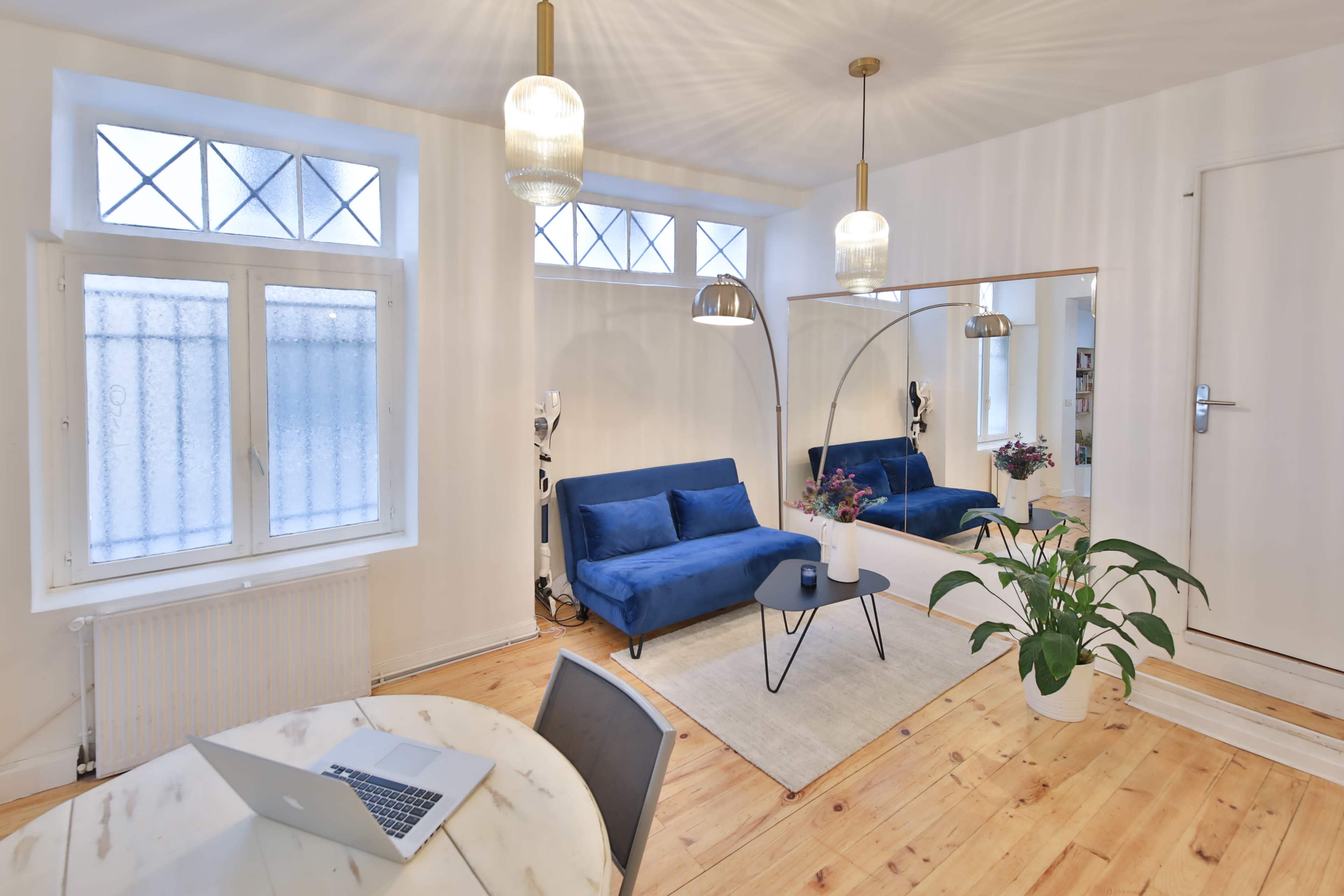 A bright living space featuring a blue sofa, a round coffee table, a large mirror, and a plant, with a dining area in the foreground and windows letting in natural light.