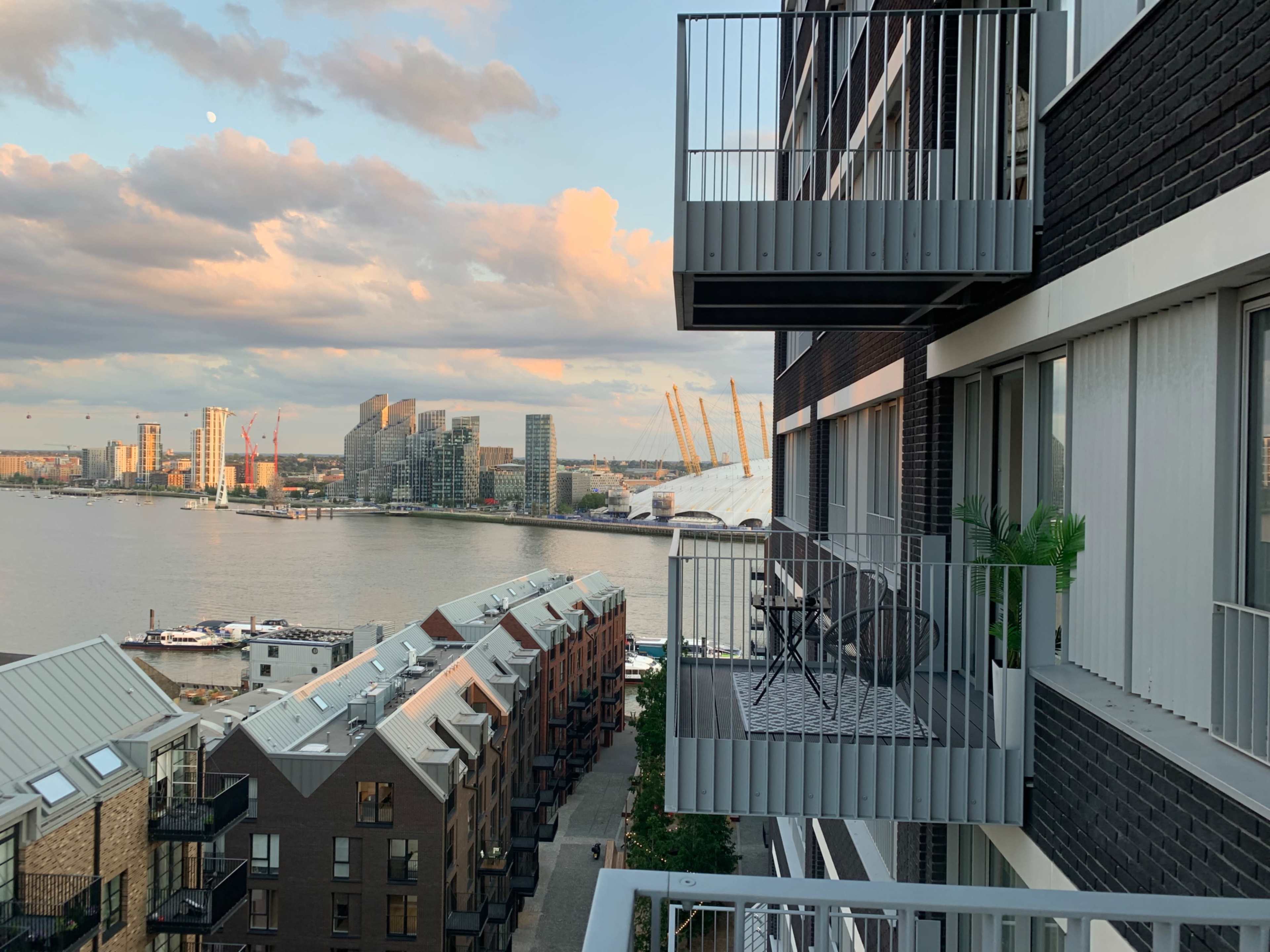 A residential building with balconies overlooking a river and modern architecture in the background during sunset.