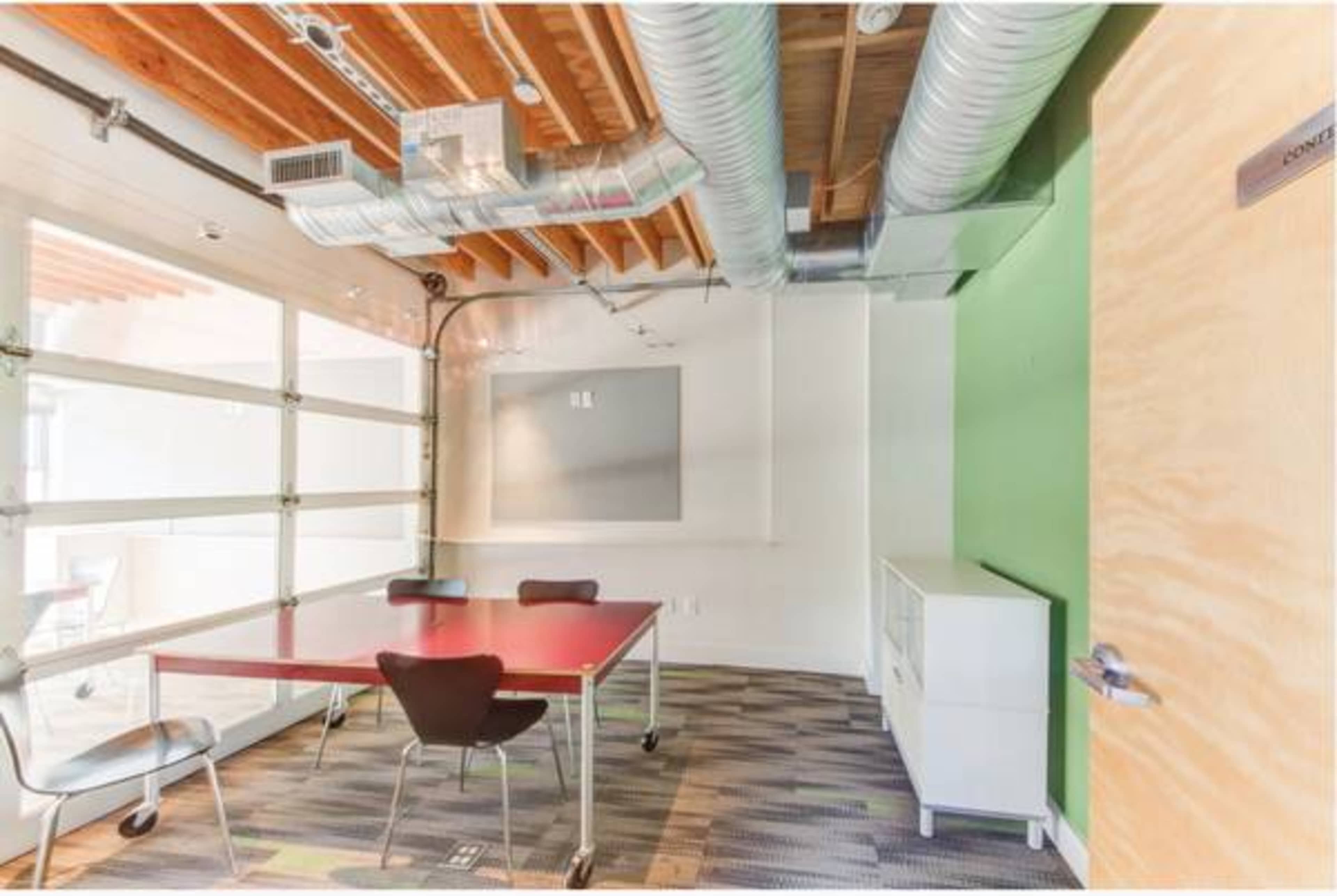 The image shows a small meeting room featuring a red table surrounded by black chairs, with exposed wooden beams and grey walls.