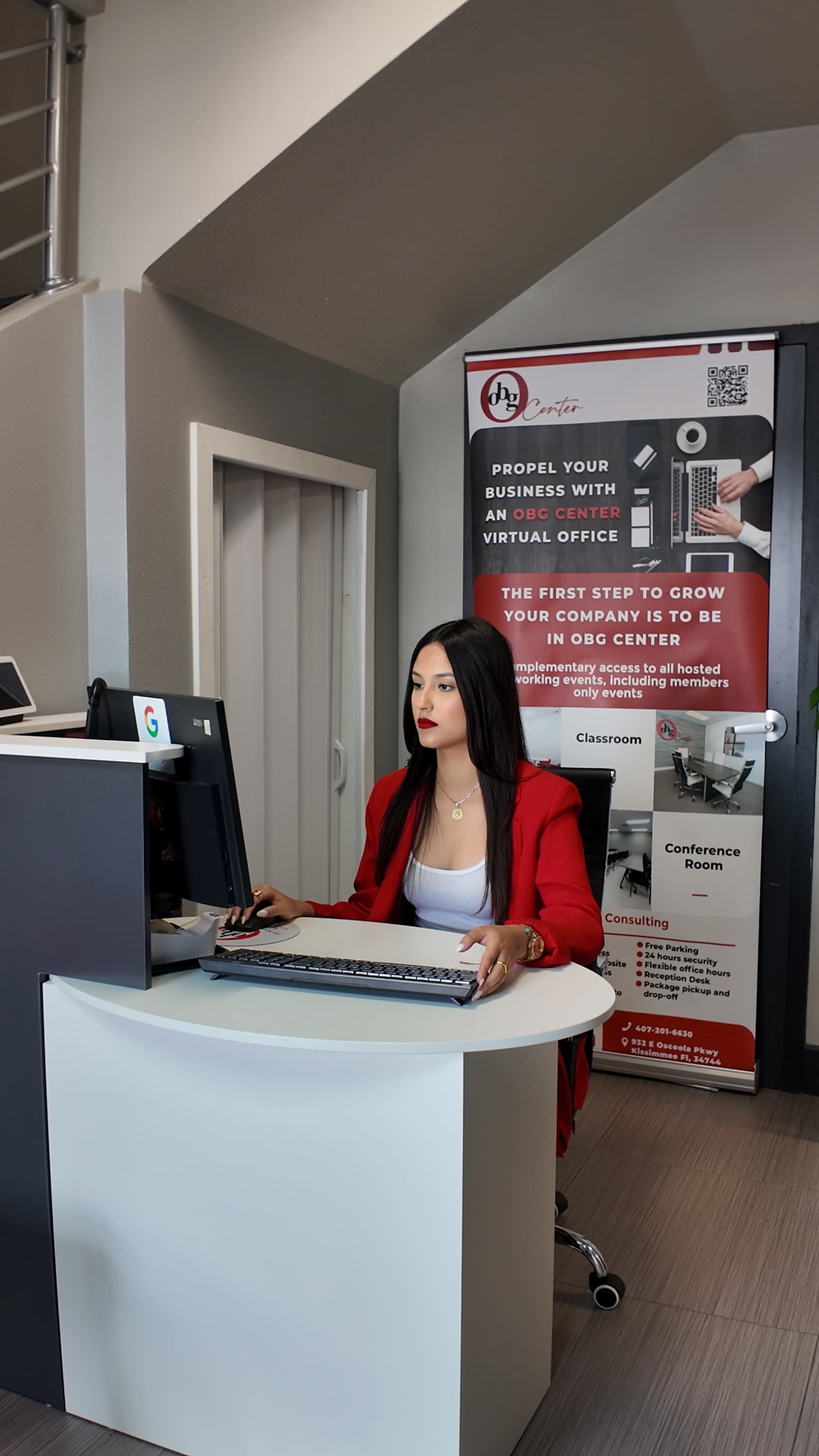 A woman in a red blazer works at a desk in an office space featuring a promotional banner for a virtual office service.