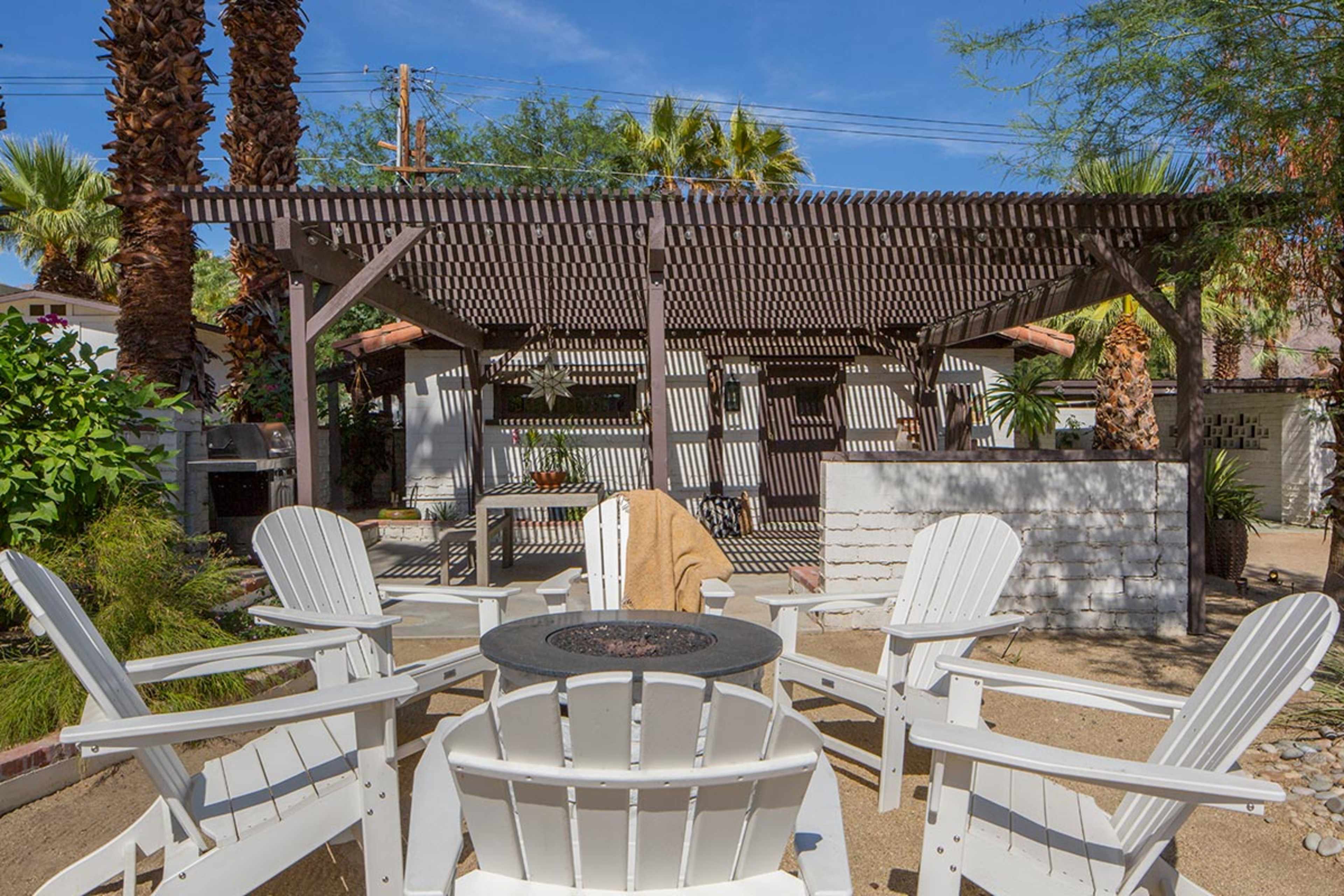 A shaded outdoor seating area features white Adirondack chairs arranged around a circular fire pit in front of a pergola and landscaped greenery.