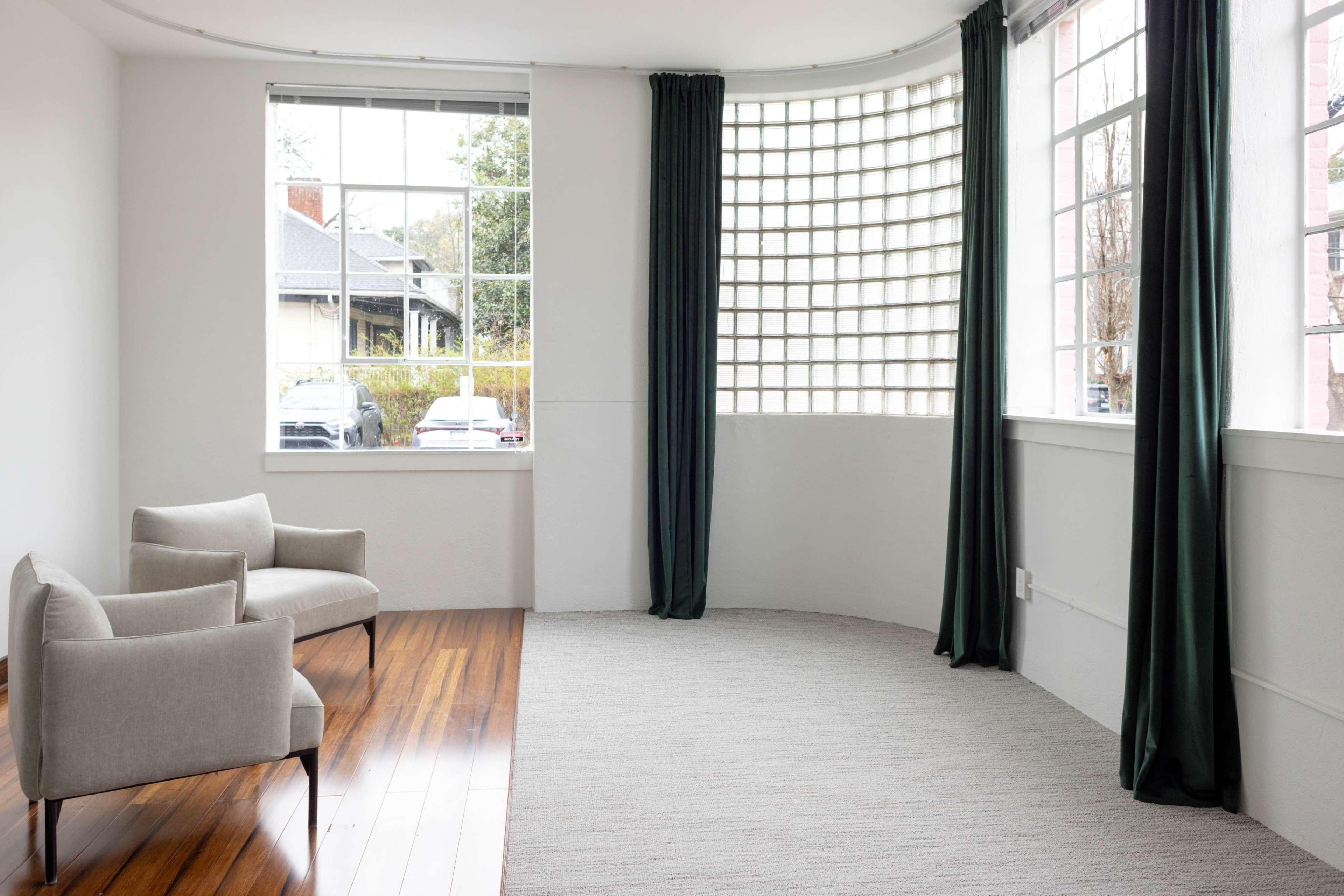 A bright room features two upholstered chairs beside large windows with green curtains, hardwood flooring, and a curved wall with frosted glass blocks.