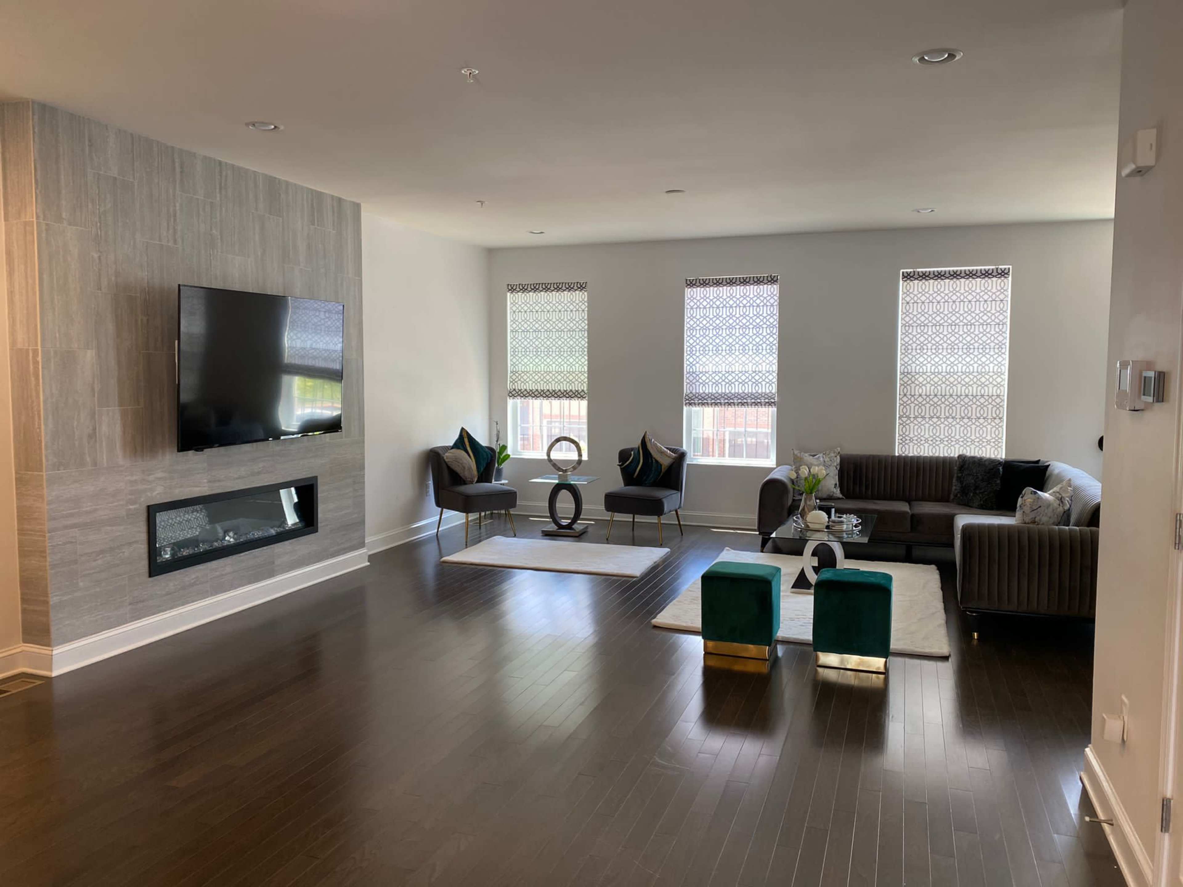 A modern living room with a large flat-screen TV mounted on a gray wall, a fireplace beneath it, and a sectional sofa surrounded by two armchairs and glass coffee tables.