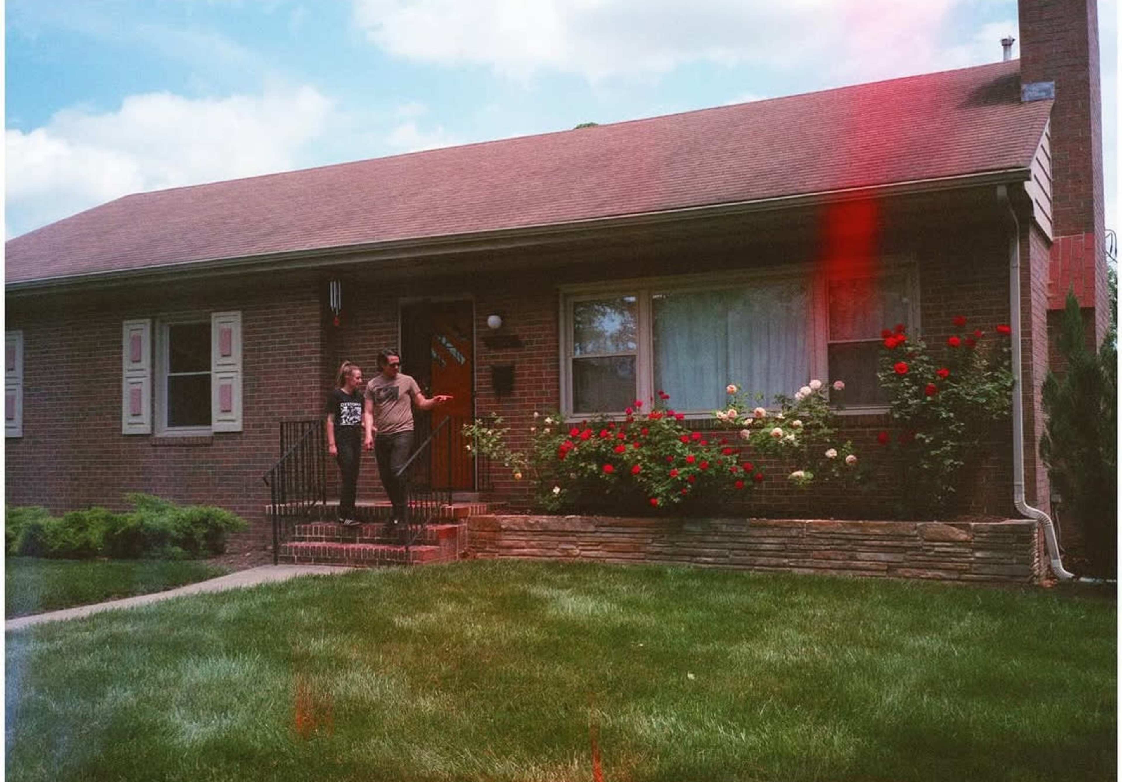 A couple stands on the front steps of a brick house, with a garden of blooming roses in front and a lawn of green grass.