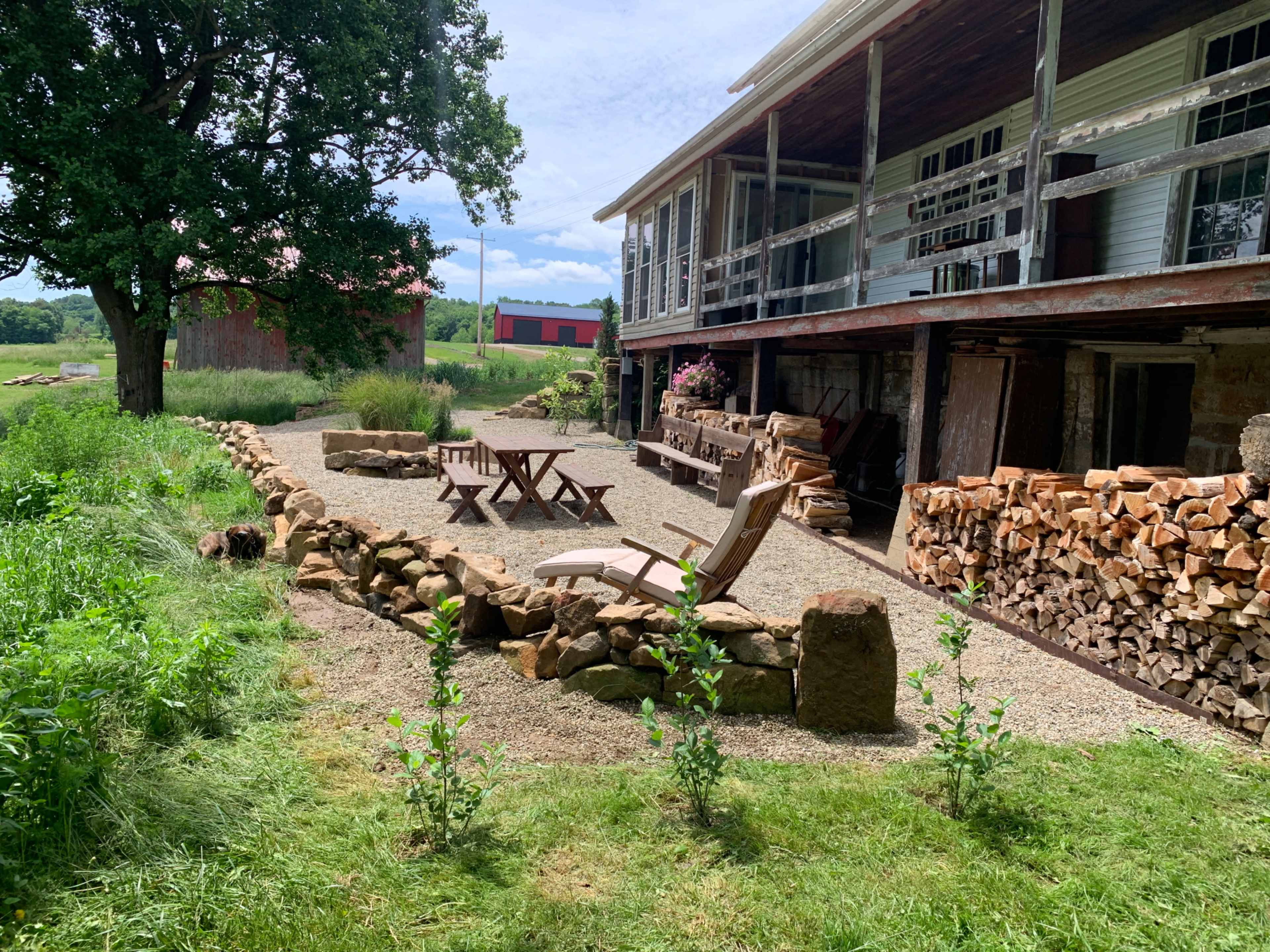 A gravel patio area with wooden seating and stacked firewood, adjacent to a multi-level house surrounded by greenery.