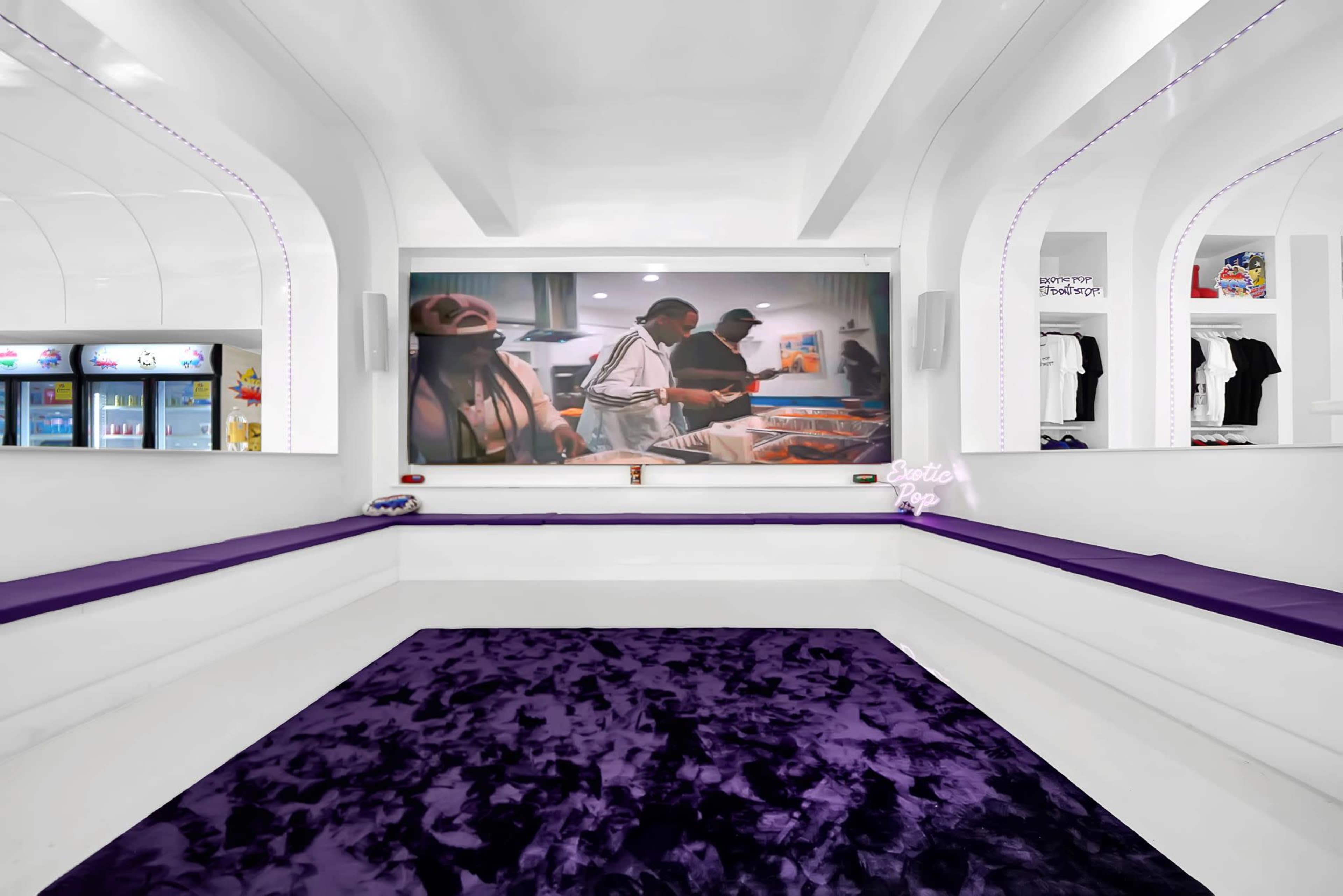 A modern, minimalist interior featuring white walls and a large screen displaying people preparing food, complemented by purple seating and a plush rug.