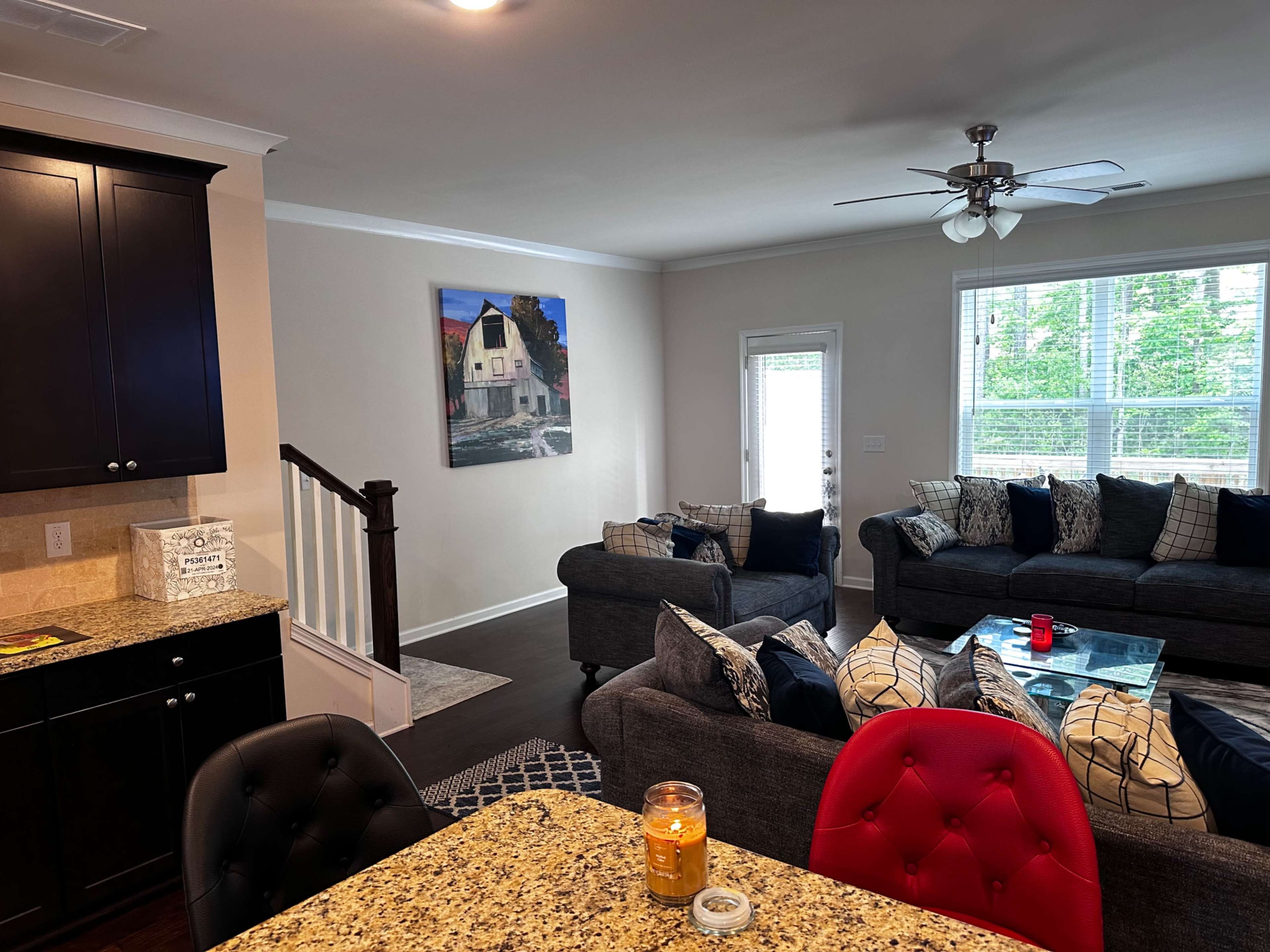 A well-lit living room with two gray couches, a coffee table, and a stairway leading to an upper level, visible from a kitchen area with dark cabinetry.