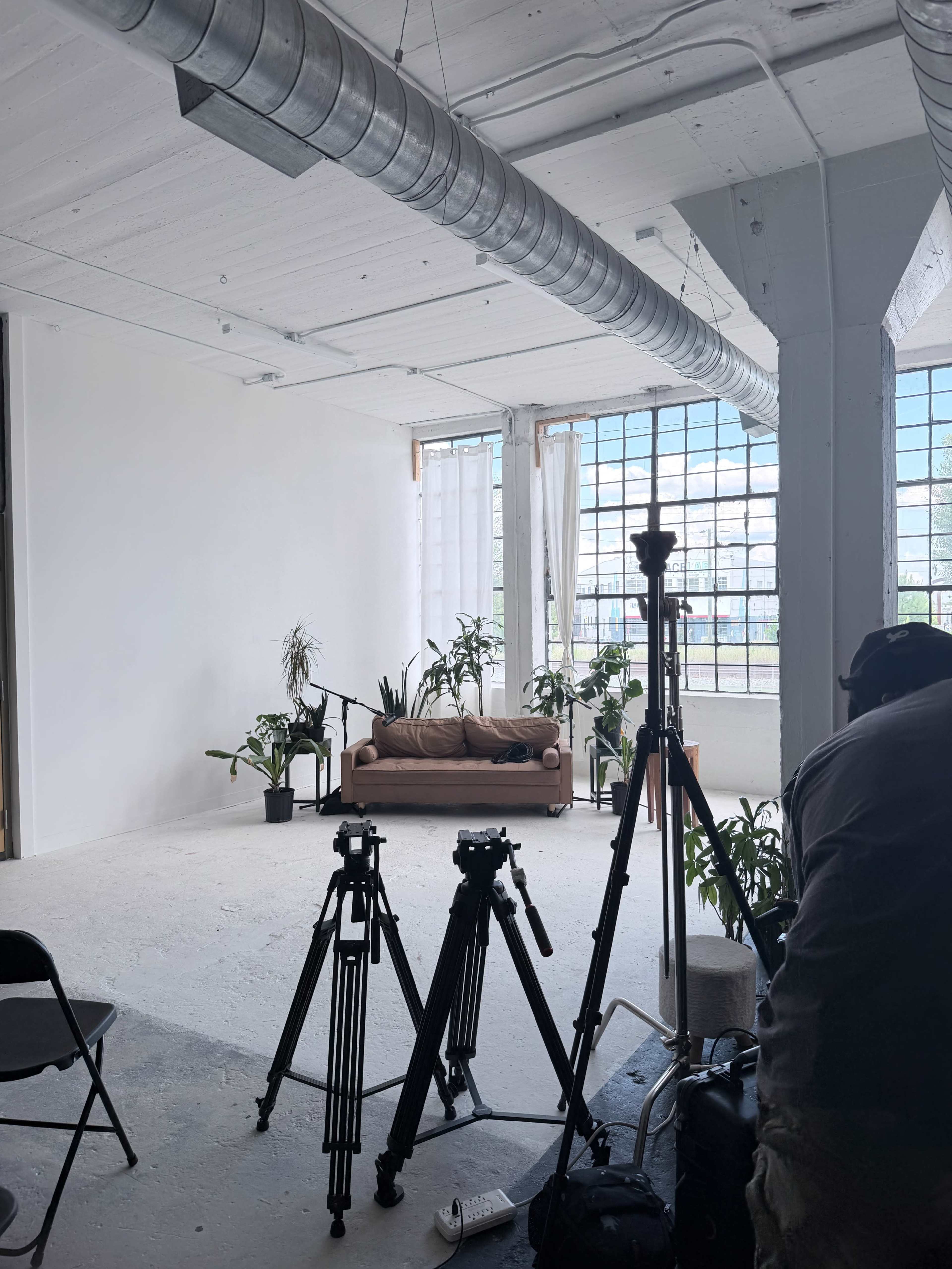 The image shows a spacious, minimalistic studio with a brown sofa, large windows, and several tripods set up for video or photography equipment, along with various indoor plants.