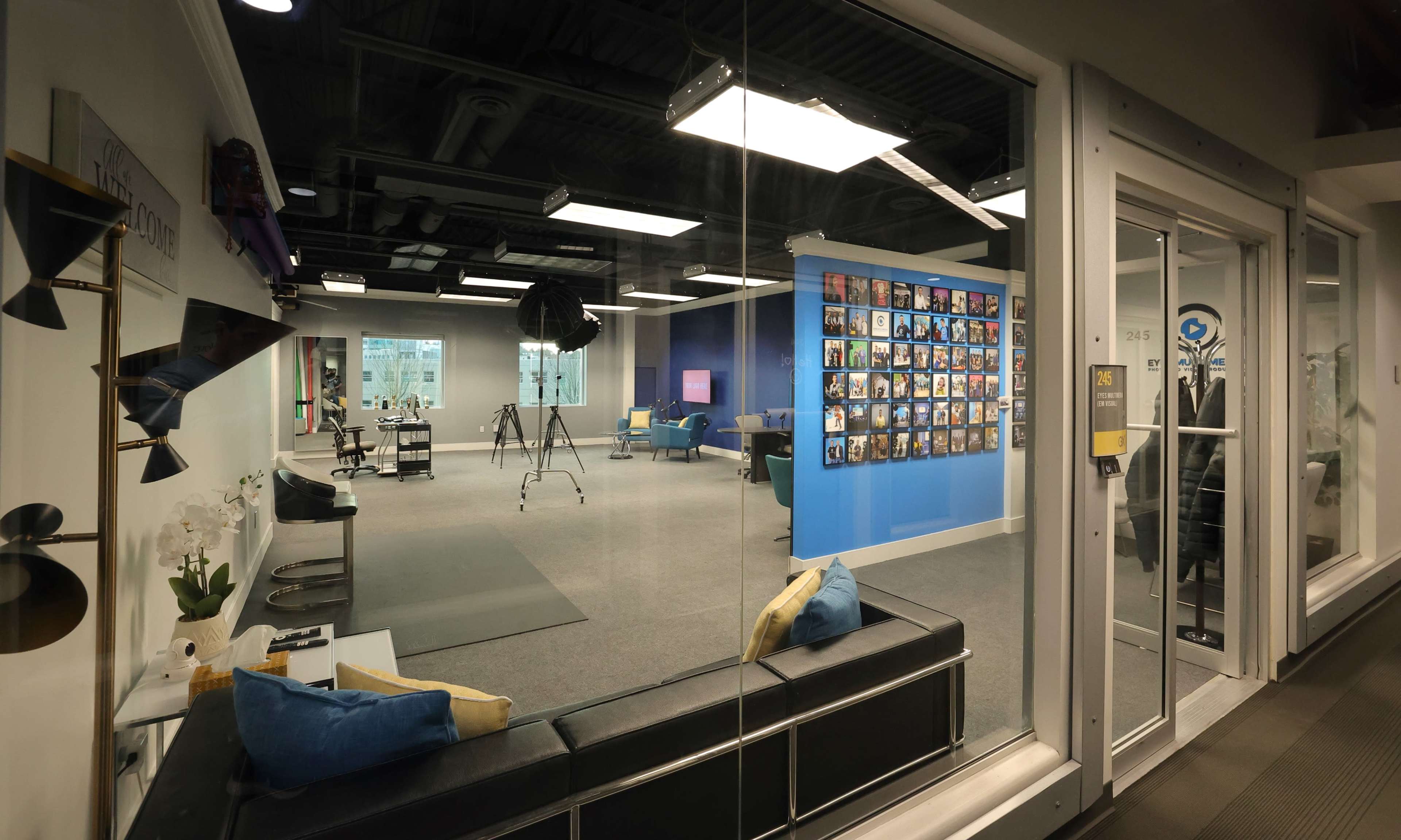 A modern office space with a seating area in front, a blue wall adorned with framed photos, and various workstations visible through large glass windows.