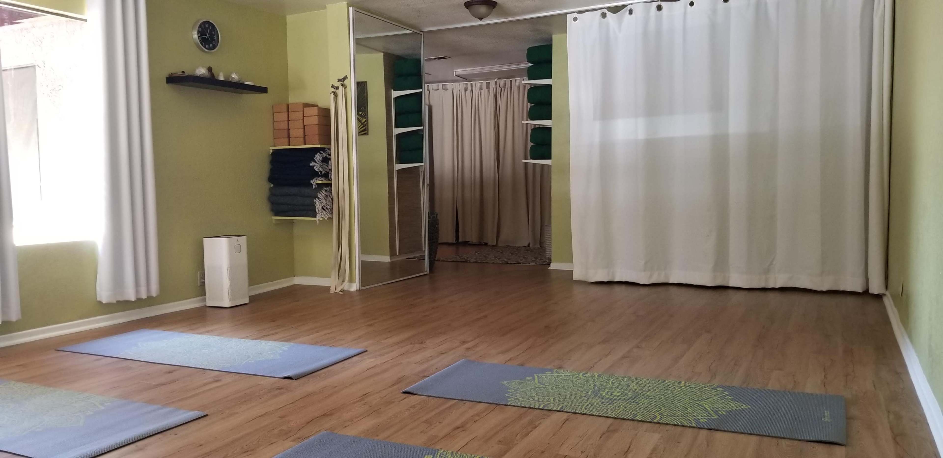 A yoga studio with wooden flooring, three yoga mats arranged in a row, and green walls adorned with shelves and curtains.
