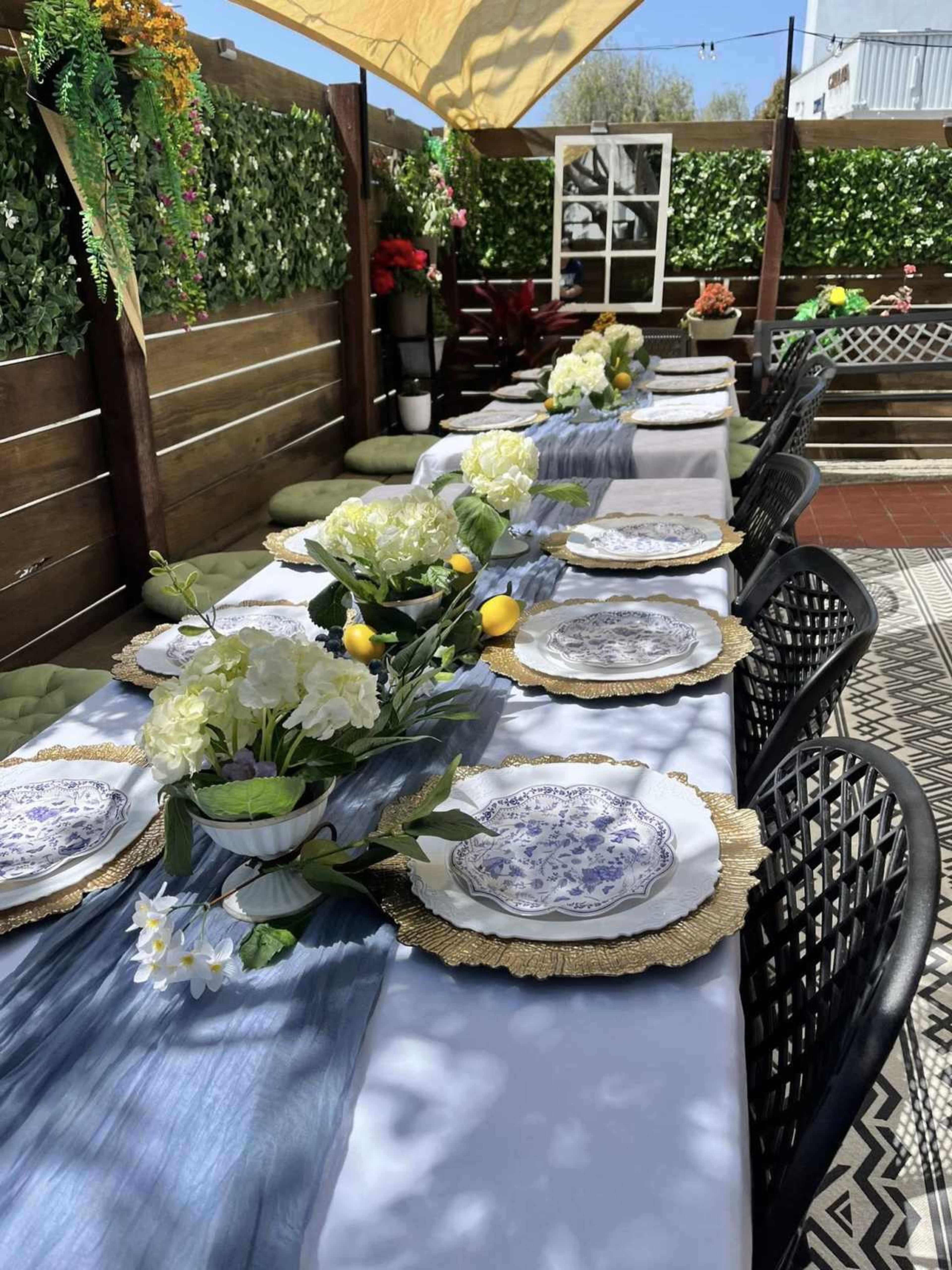 The image shows a decorated outdoor dining area with a long table set with floral centerpieces, plates, and a blue table runner under a shaded canopy.