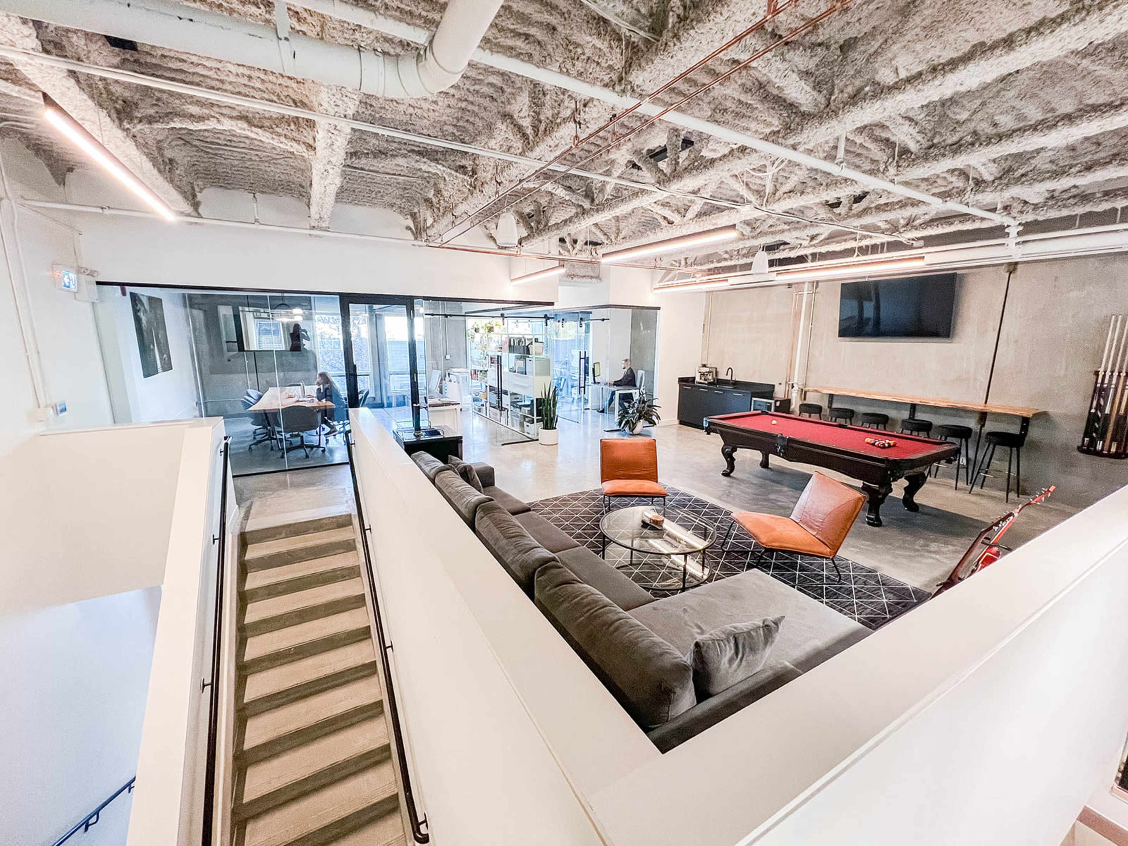 The image shows a spacious indoor lounge area with a pool table, a sofa, and two chairs, overlooking an adjacent glass-enclosed office space.
