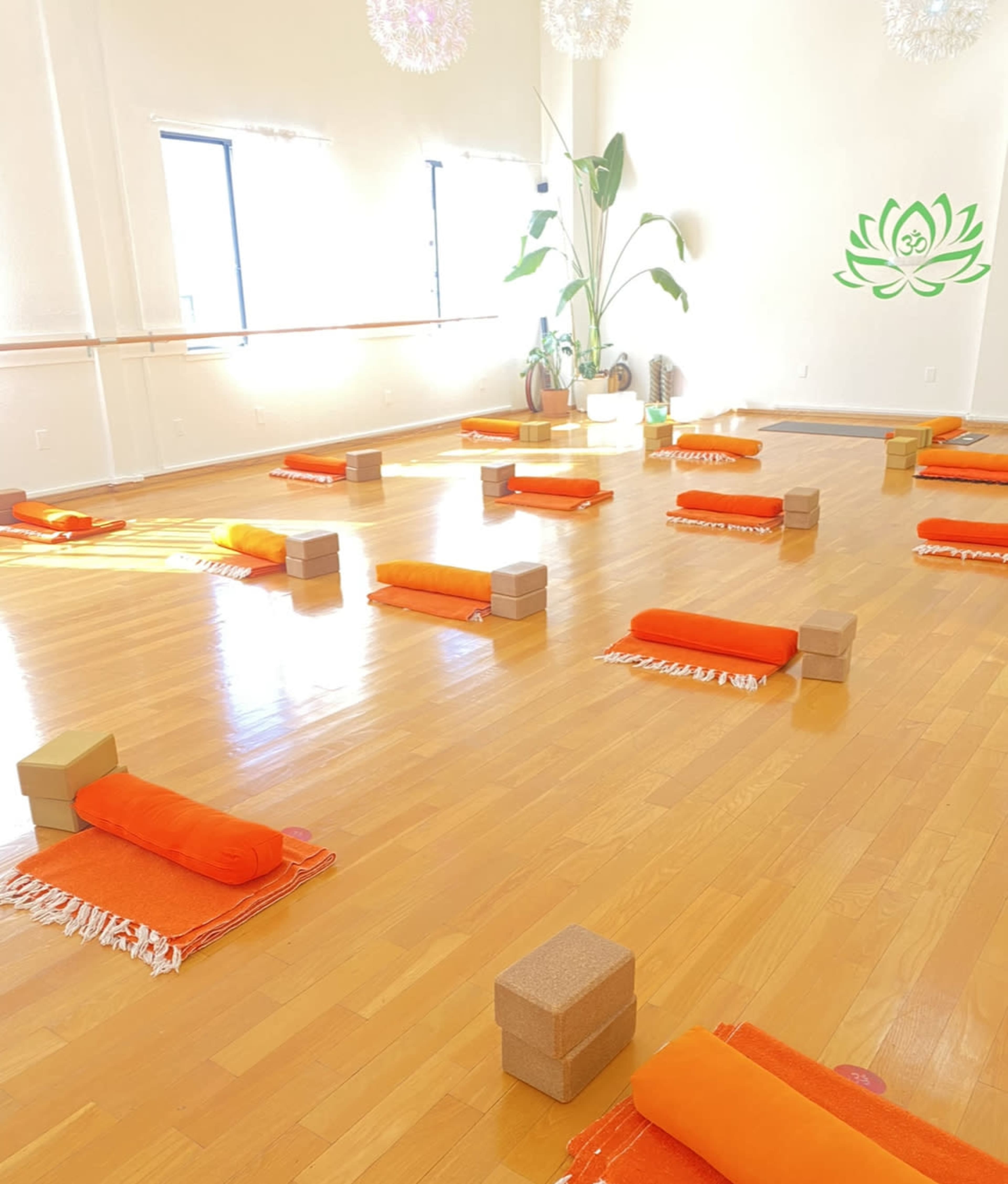A yoga studio with wooden floors arranged with orange mats and props, illuminated by natural light from large windows.