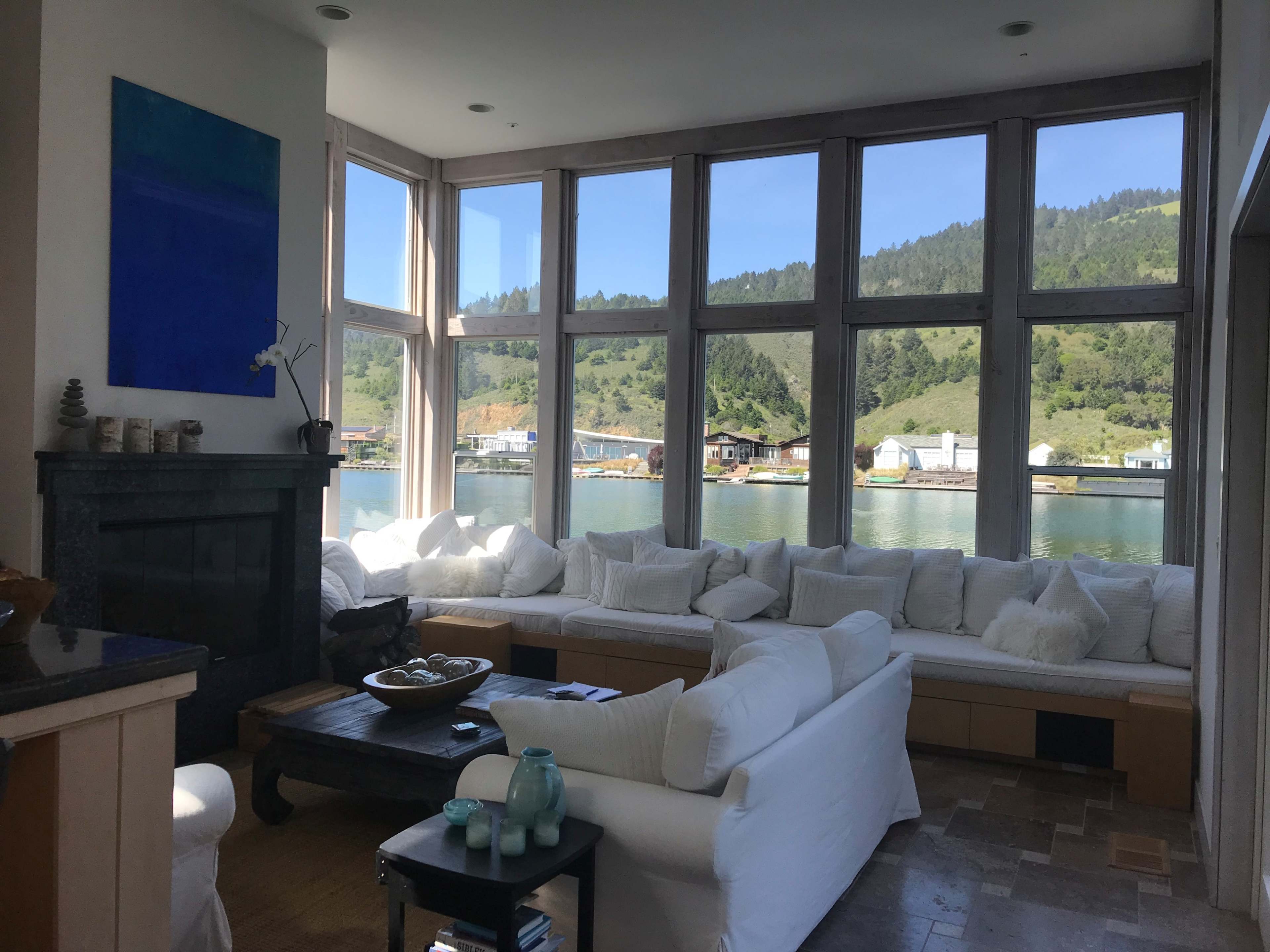 A modern living room with large windows offering a view of a calm waterfront and green hills.
