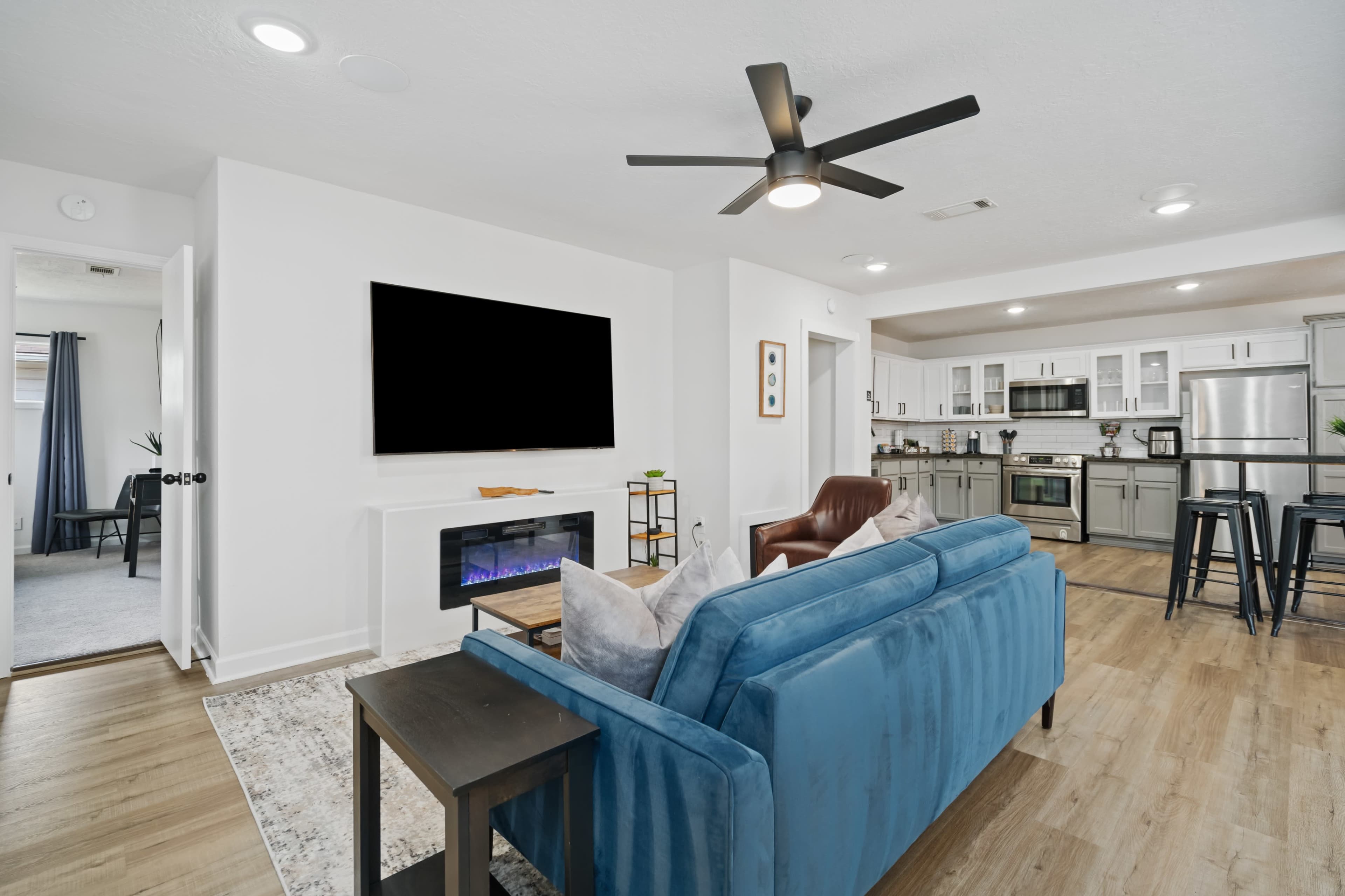 A modern living room features a blue sofa, a wall-mounted television, and an electric fireplace, with an open layout leading to a kitchen area.