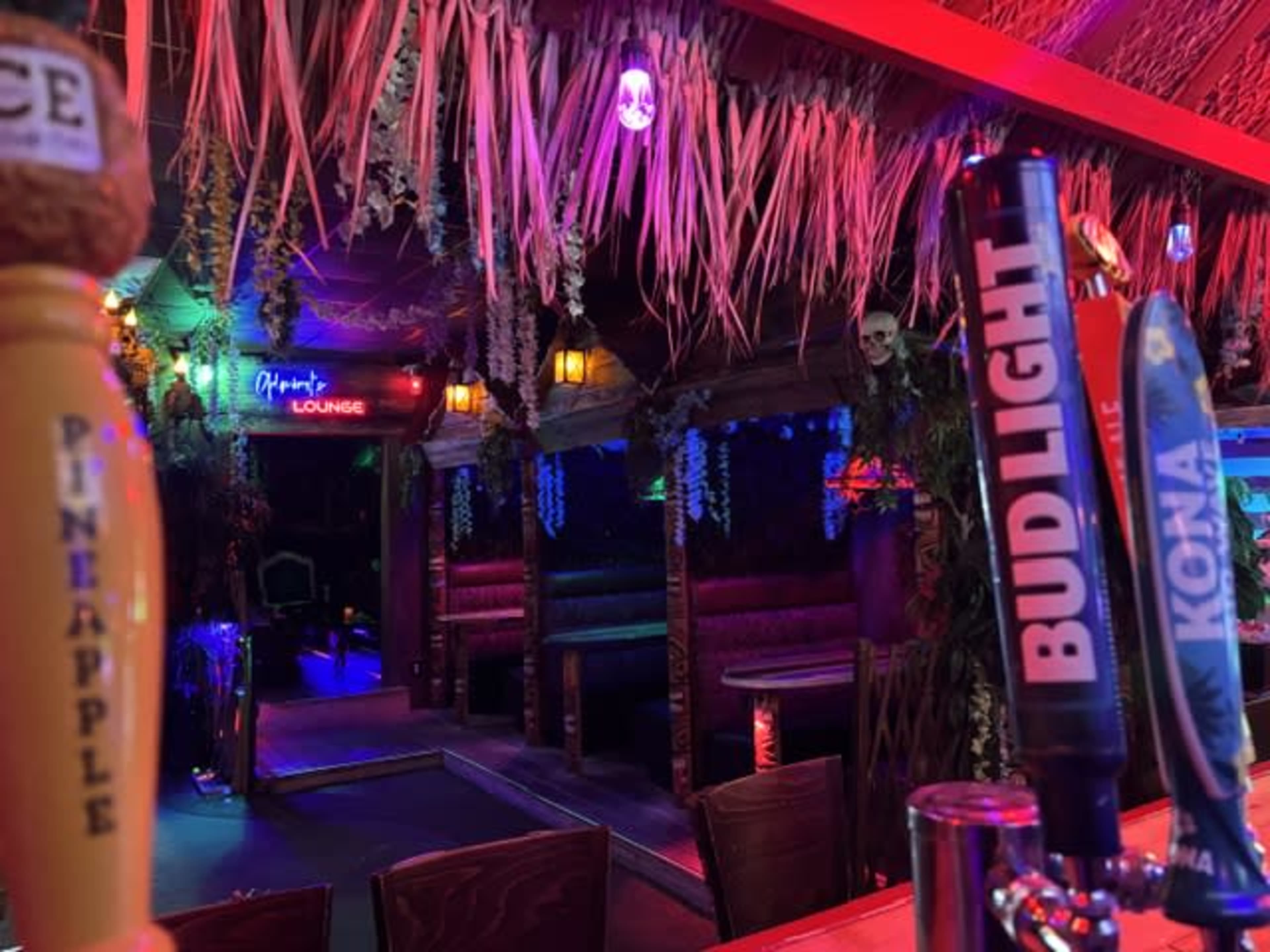 A bar area features several beer taps with vibrant neon lighting and tiki-style decor, including hanging plants and dimmed seating areas.