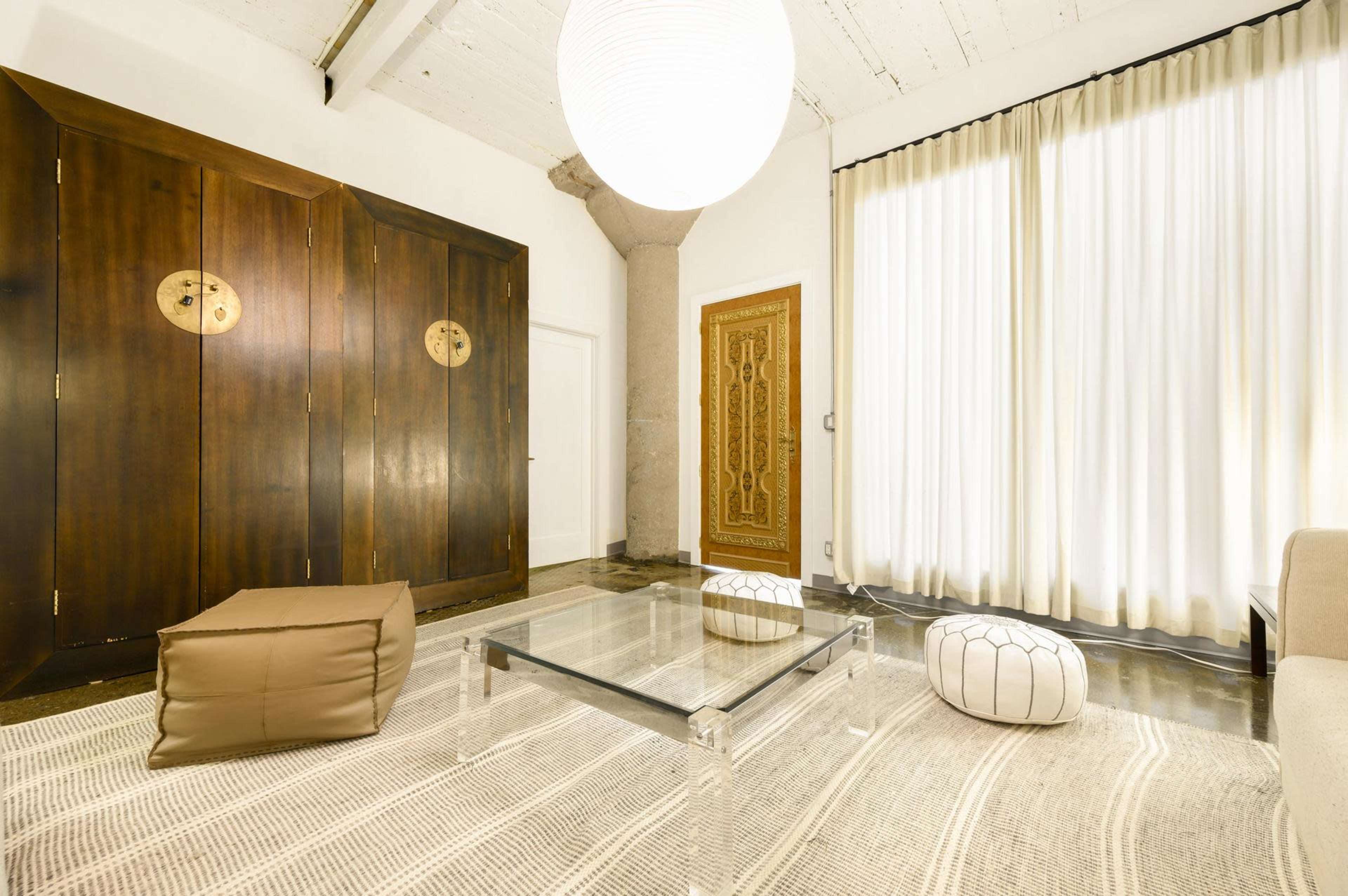 The image shows a modern living room featuring a glass coffee table, an area rug, and two decorative poufs, with large sheer curtains and wooden cabinets in the background.