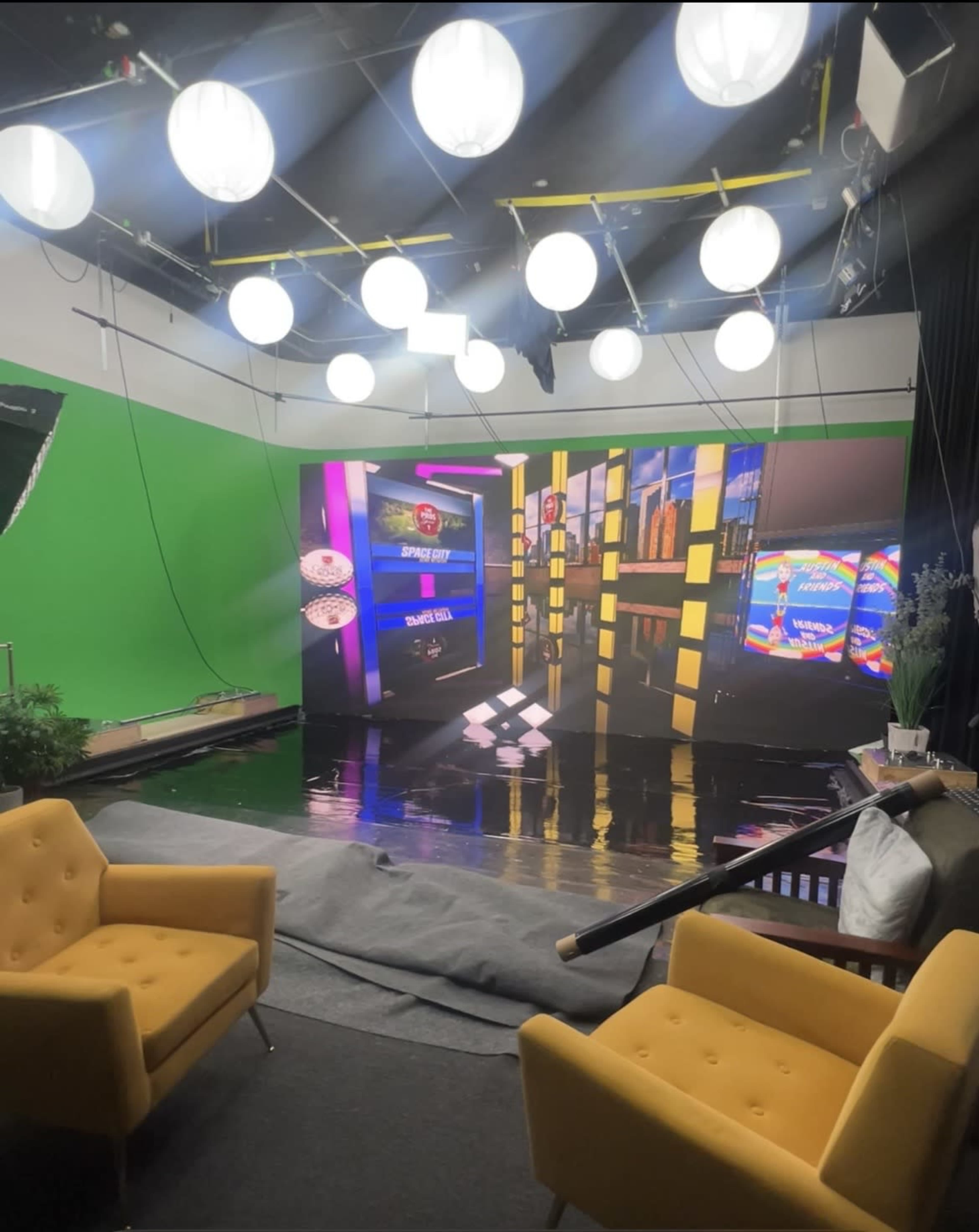 The image shows a television studio setup with green screens, large circular lights overhead, and a modern stage featuring screens and a checkered floor.