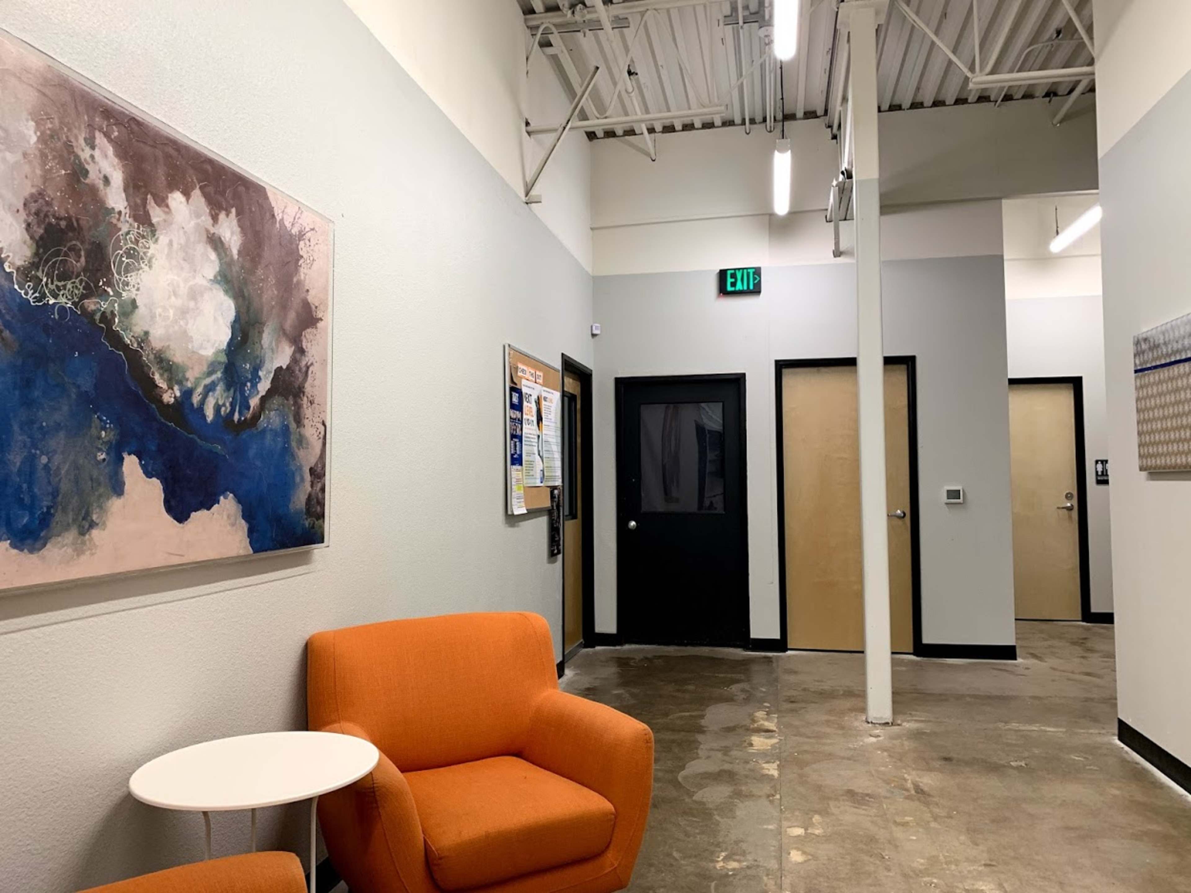 A hallway with a seating area featuring two orange chairs, an abstract painting on the wall, and doorways leading to other rooms.