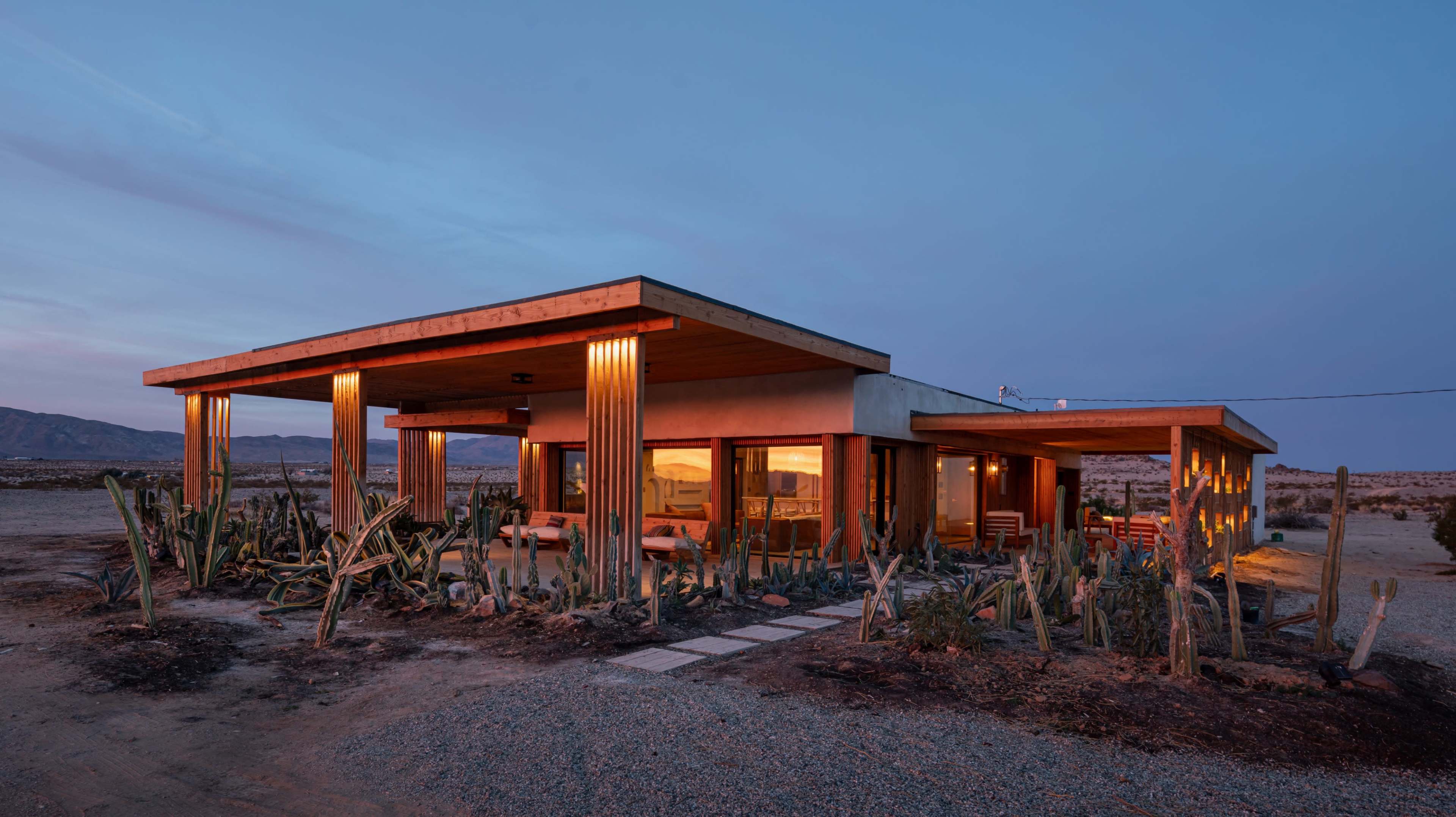 The image shows a modern house with large windows, wooden accents, and surrounding desert vegetation, set against a twilight sky.