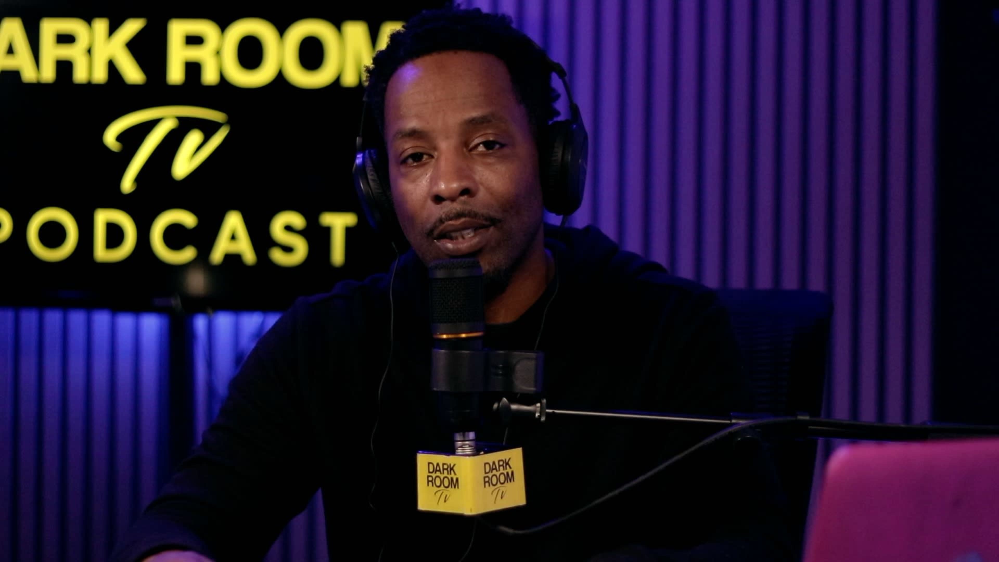 A man wearing headphones speaks into a microphone in a podcast studio with "DARK ROOM TV PODCAST" displayed in the background.