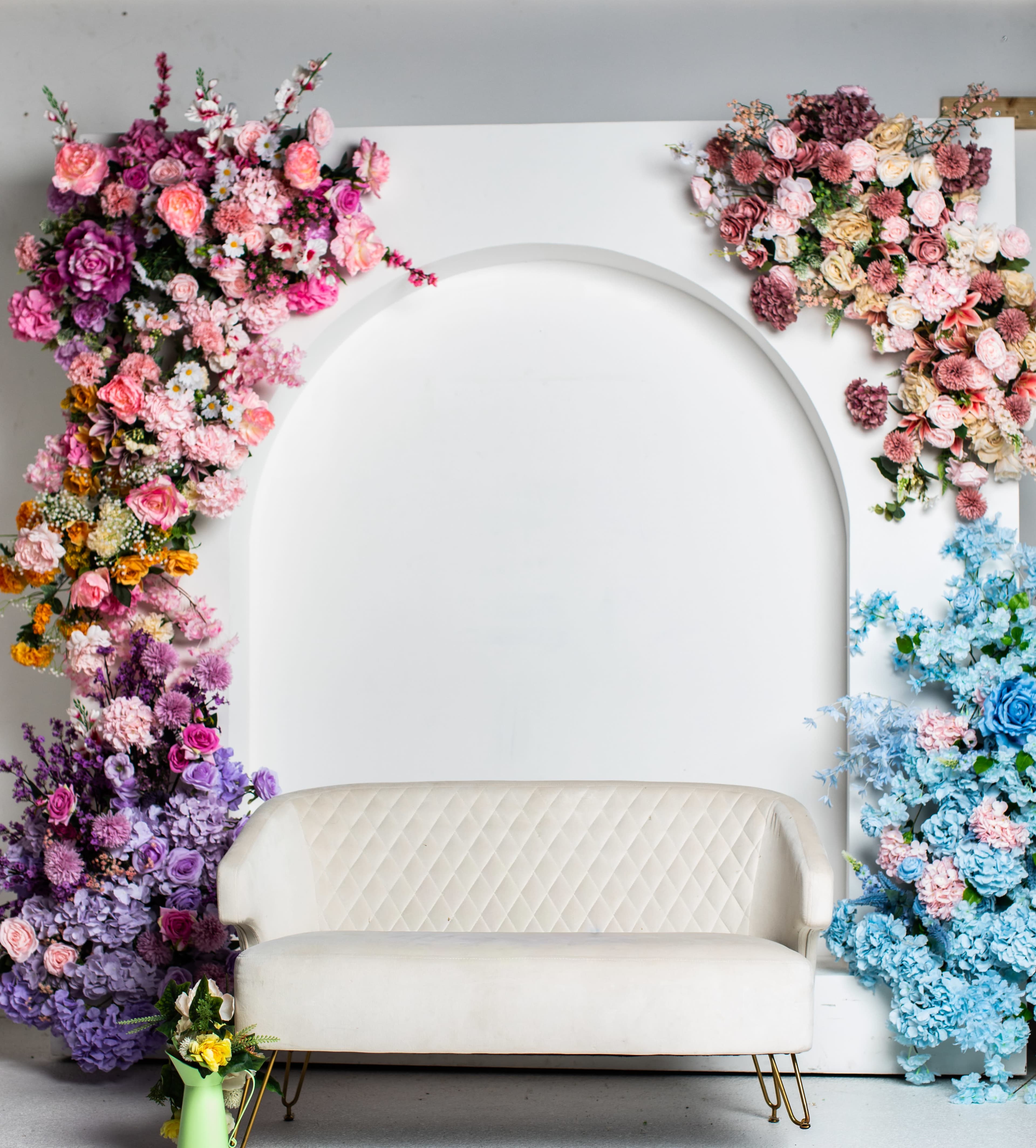 The image features a white, quilted loveseat set against a backdrop adorned with colorful floral arrangements on either side of an archway.