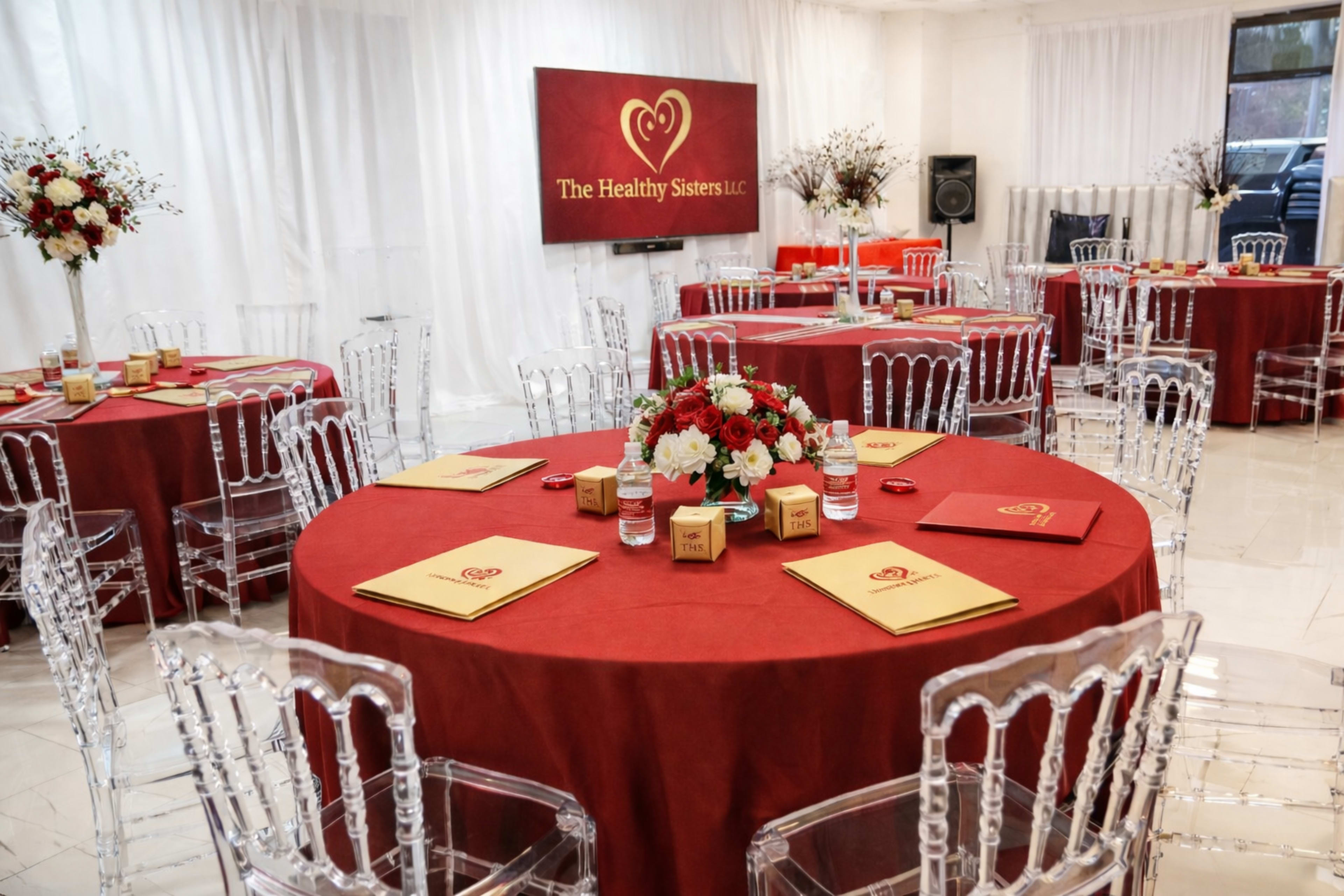 The image shows a banquet room set up with round tables covered in red tablecloths, each adorned with floral centerpieces and surrounded by clear acrylic chairs, under white draped walls and a branded display.