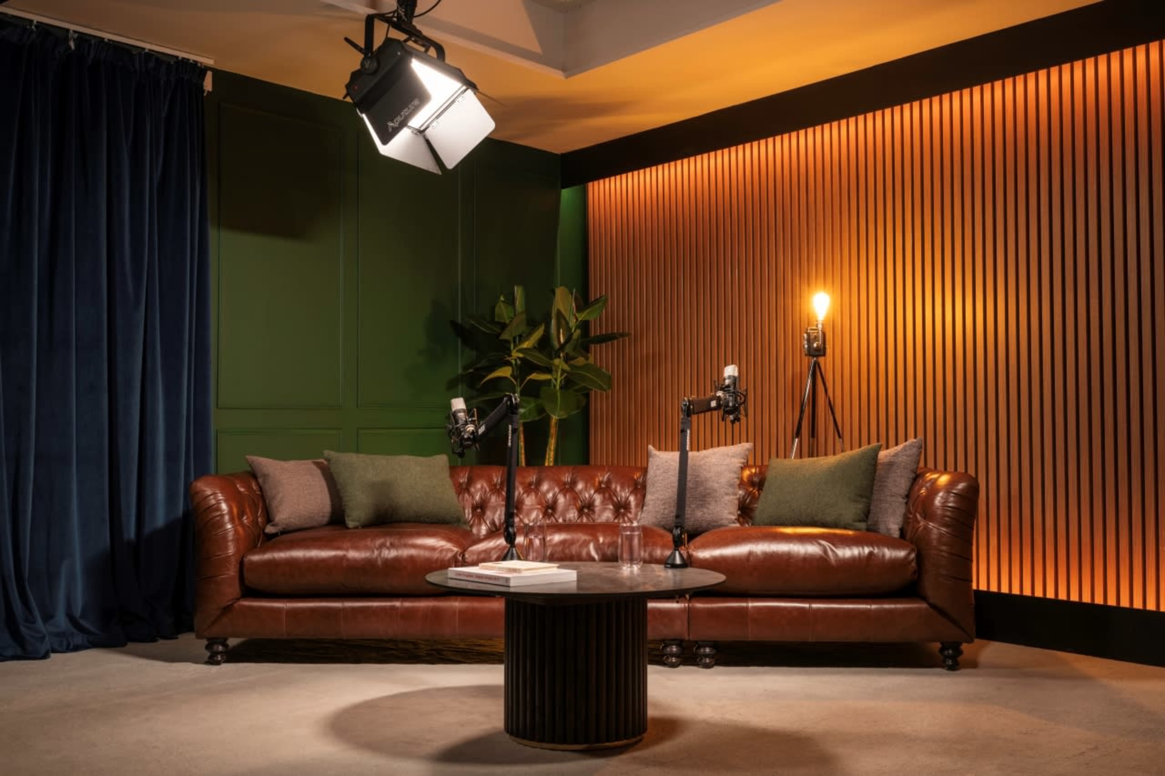 A leather chesterfield sofa with green pillows is positioned in a room featuring warm backlit wood paneling and a potted plant, with microphones mounted on stands.