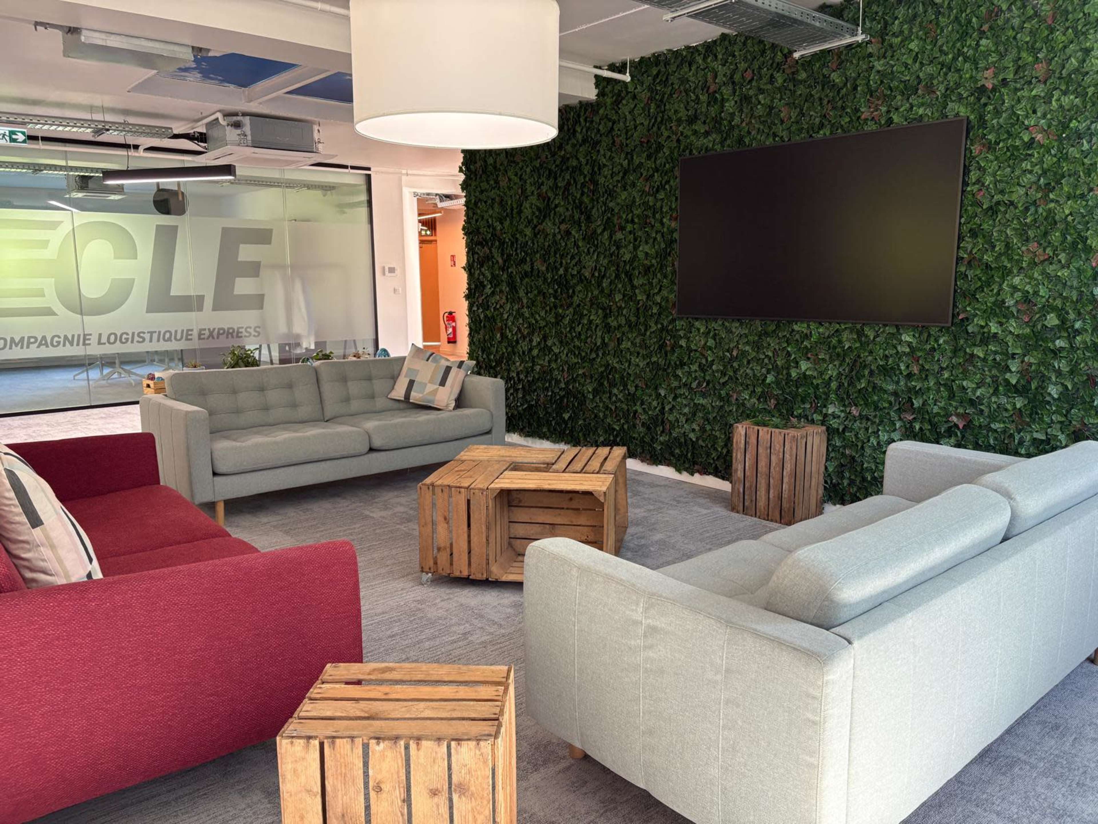 The scene shows a modern lounge area with three sofas, a coffee table made of wooden crates, and a large flat-screen TV mounted on a green wall covered in artificial foliage.
