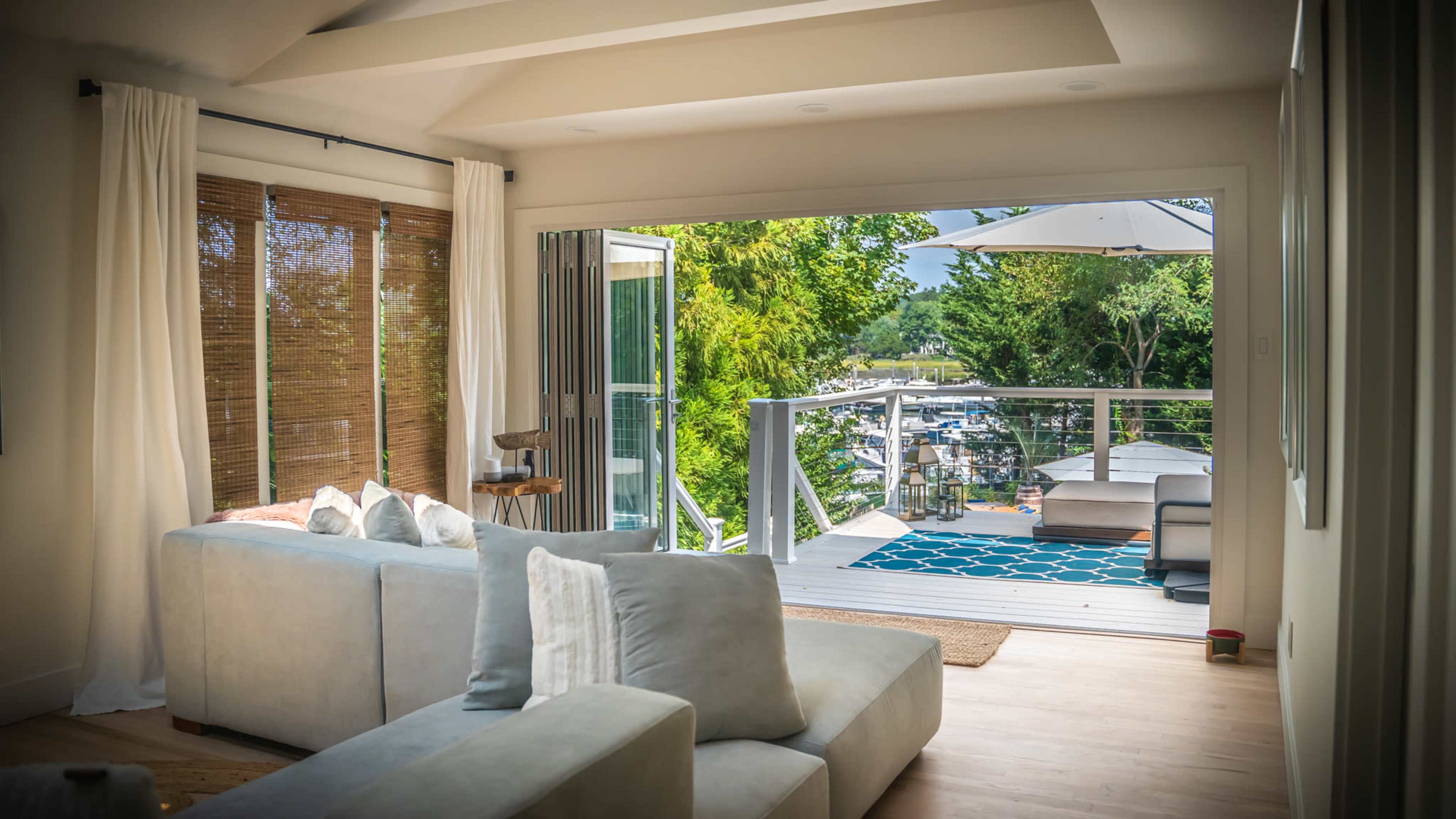 A living room with large windows opening onto a deck overlooking a marina, featuring a sectional sofa and a rug.