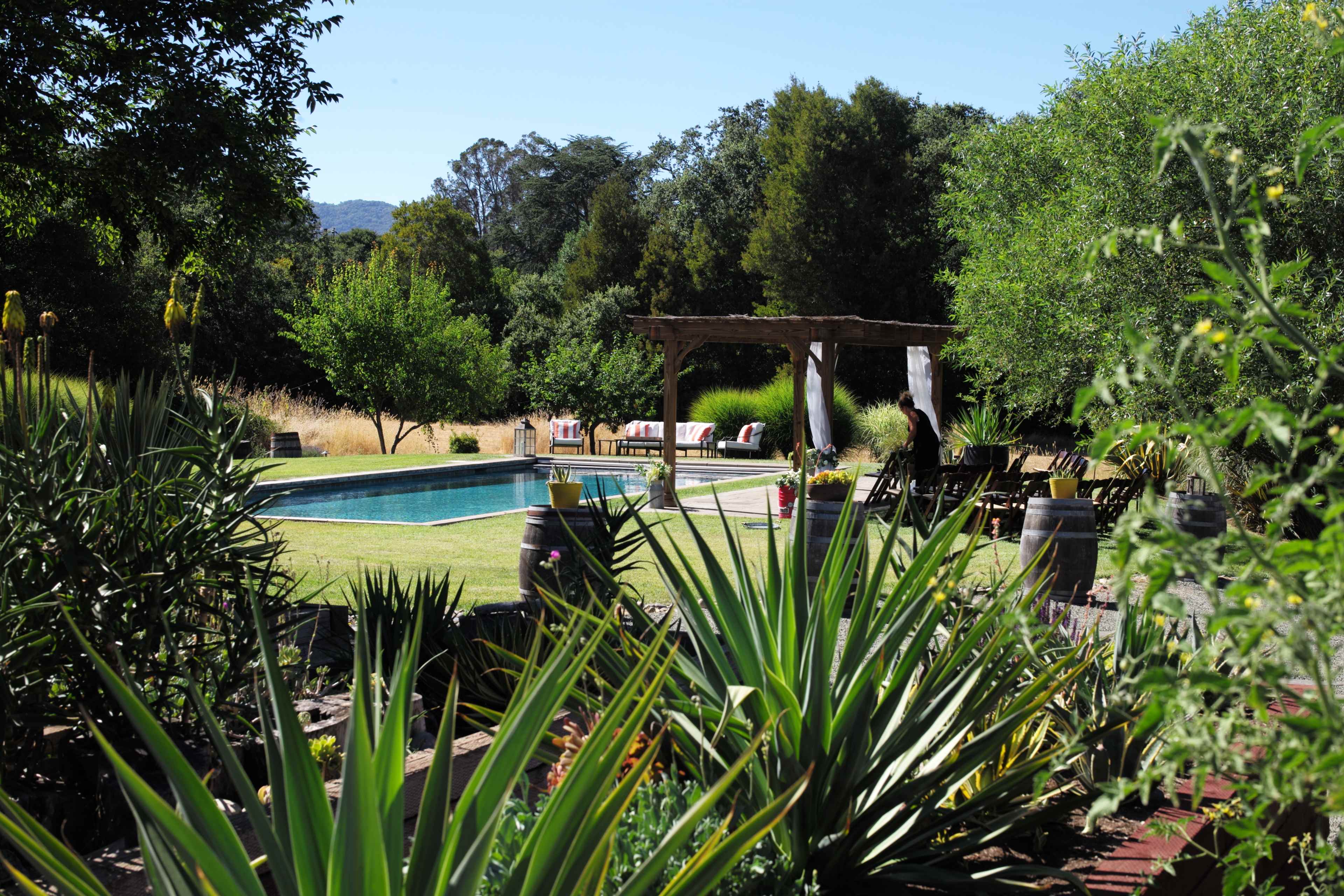A backyard pool surrounded by lush greenery and seating areas under a wooden pergola.
