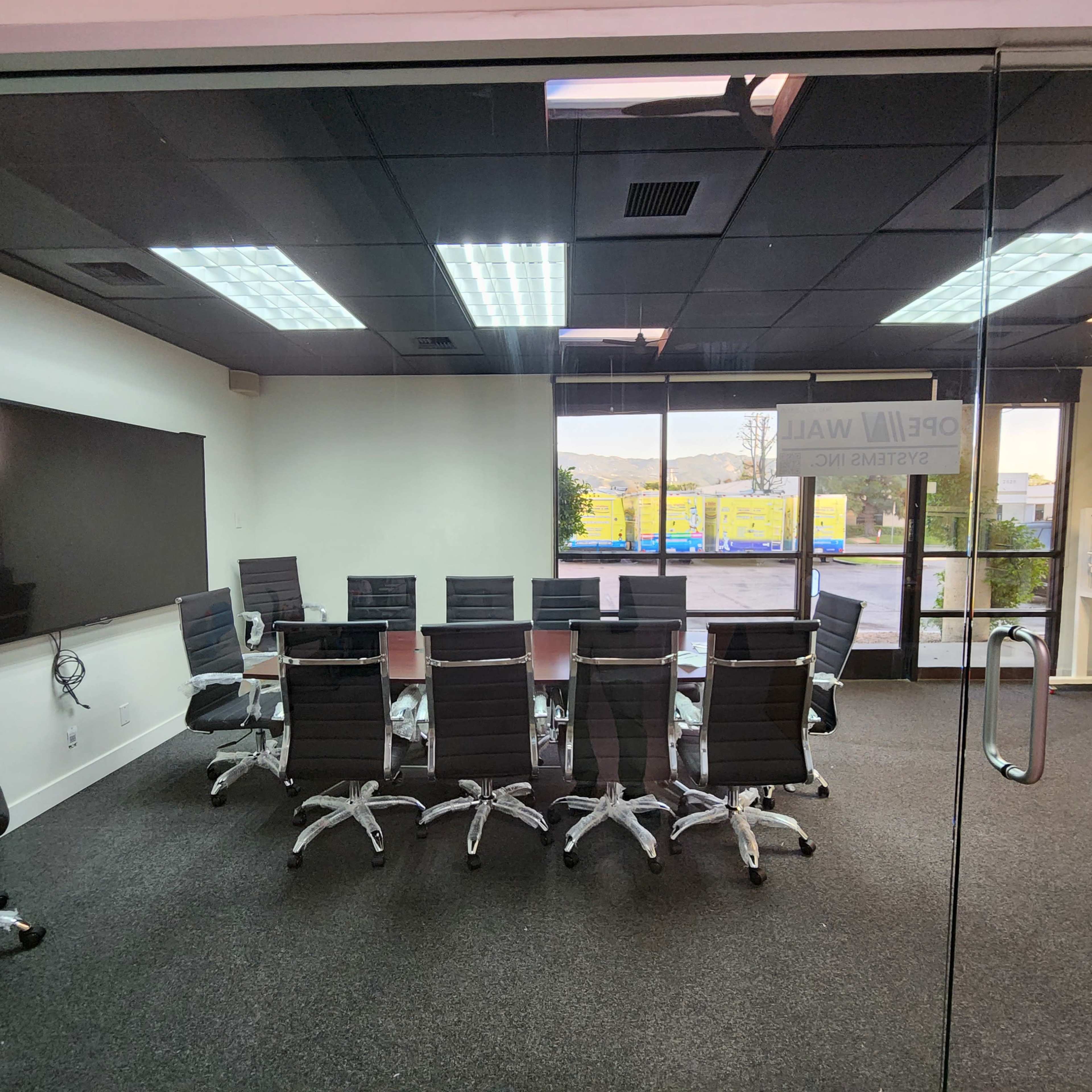 A glass-walled conference room features a large, oval table surrounded by rolling chairs and a wall-mounted display panel.