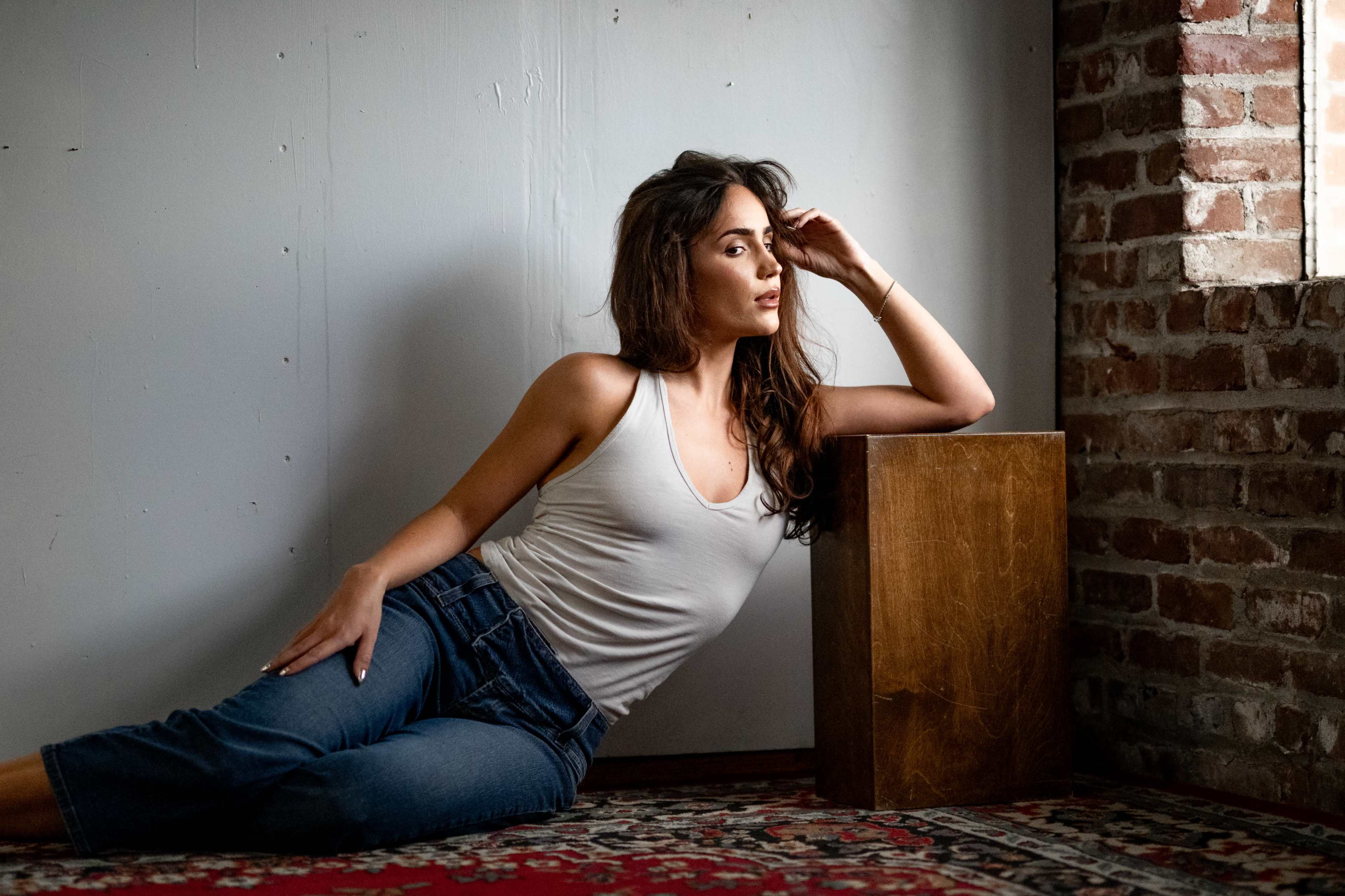 A woman in a tank top is leaning against a wooden block, sitting on a patterned rug in front of a brick wall.