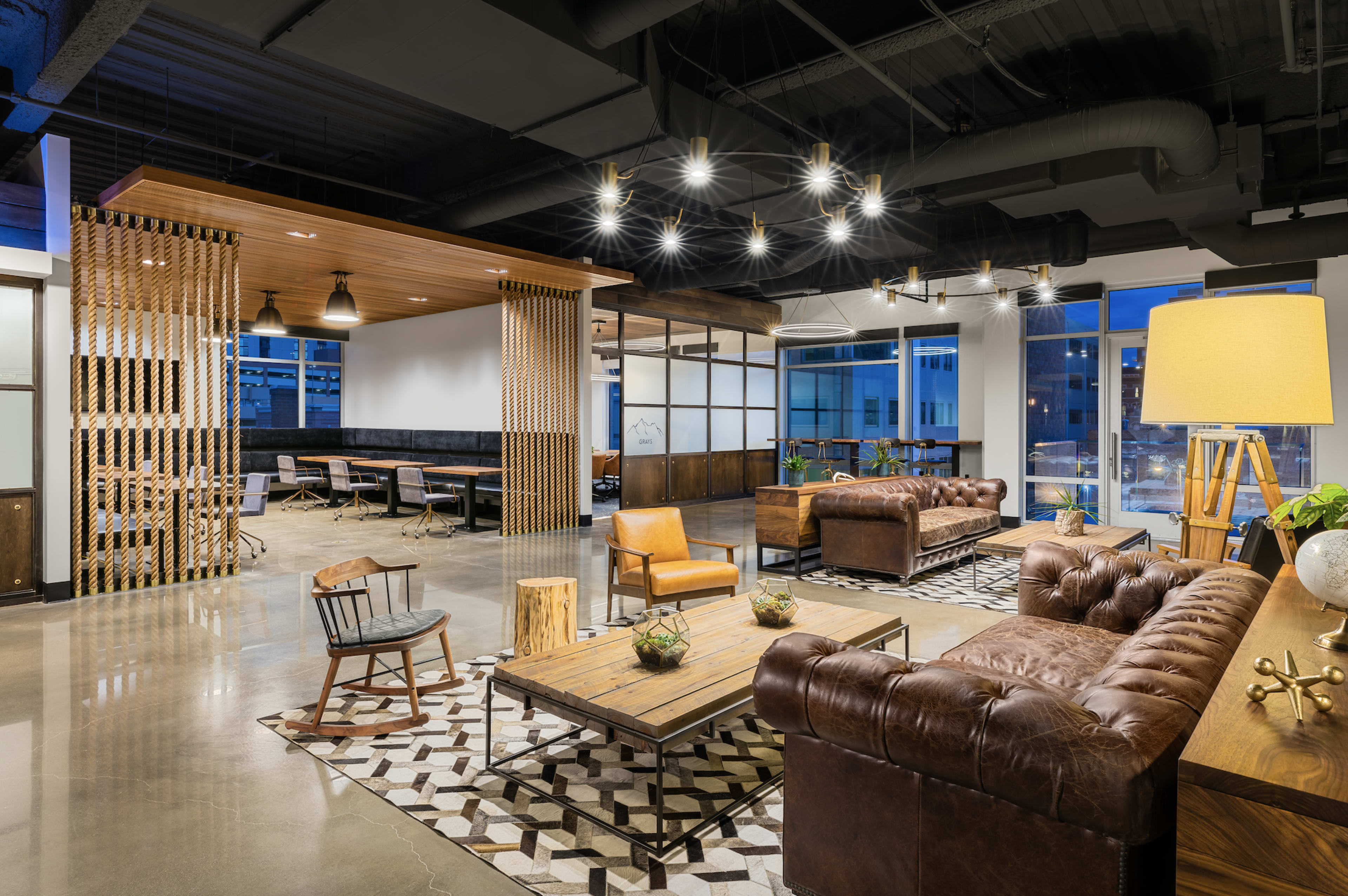 The image shows a modern office space with a mix of lounge seating, wooden tables, and a collaborative work area, illuminated by pendant lights and large windows.