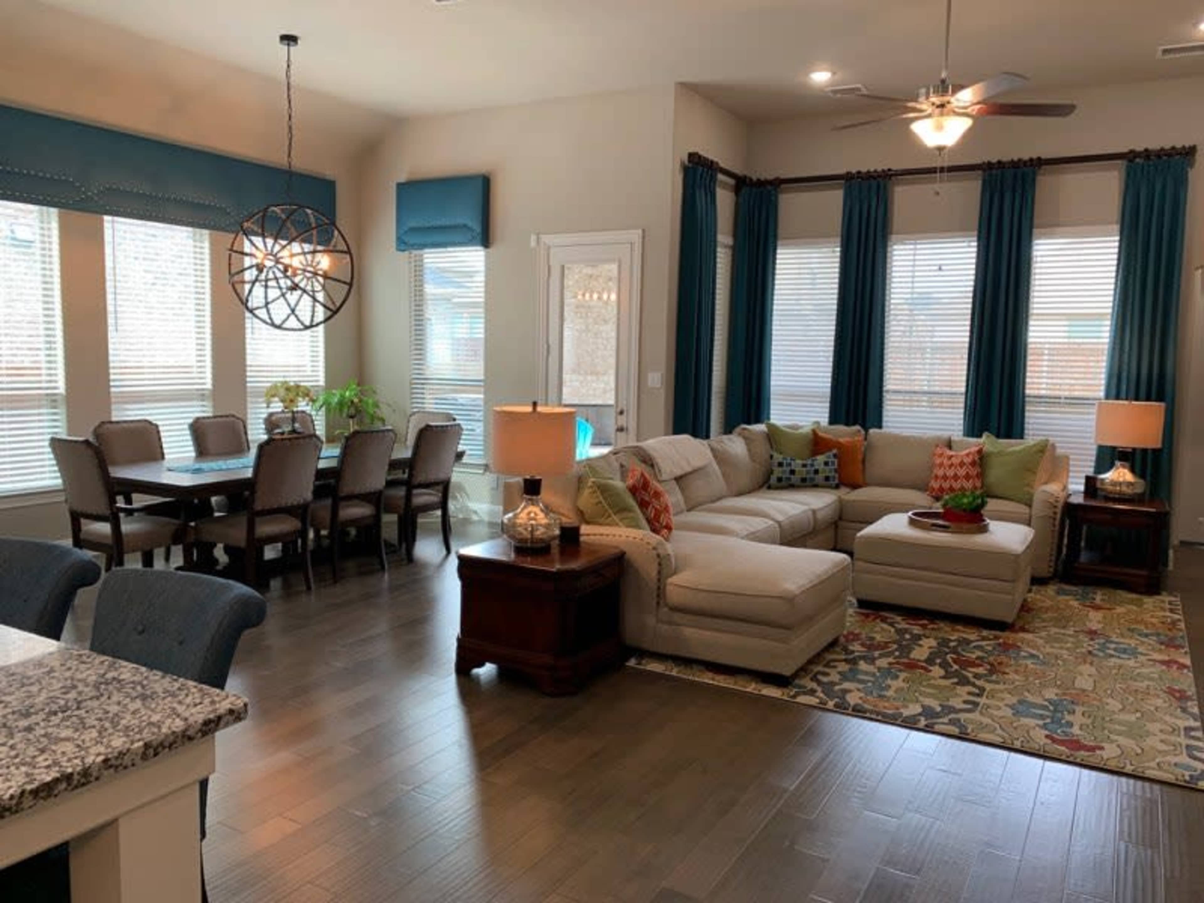 A living room and dining area featuring a sectional sofa, a dining table with chairs, and large windows with blue curtains.