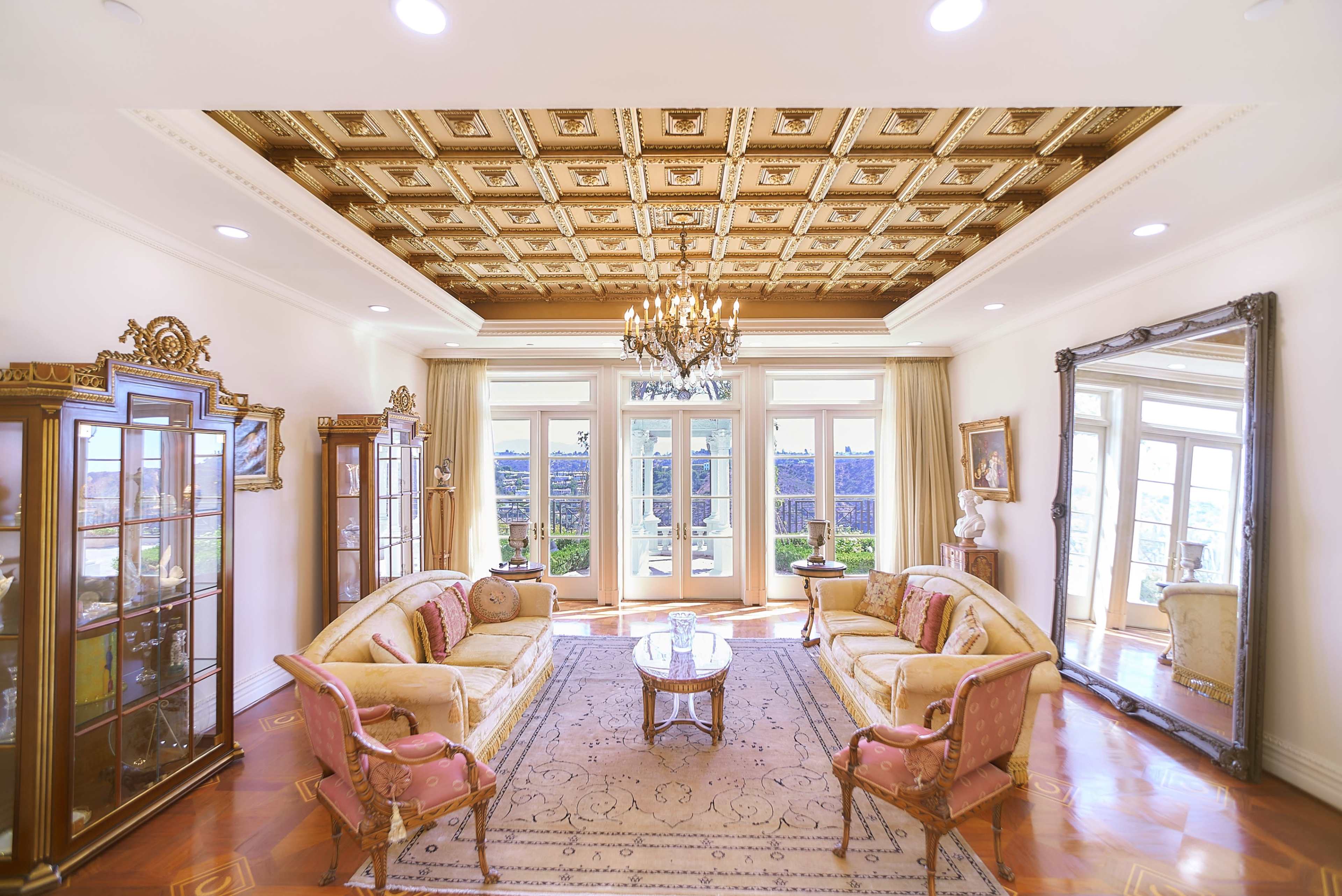 A spacious living room furnished with two sofas, a coffee table, and a large mirror, featuring ornate details and large windows that open to a view outside.