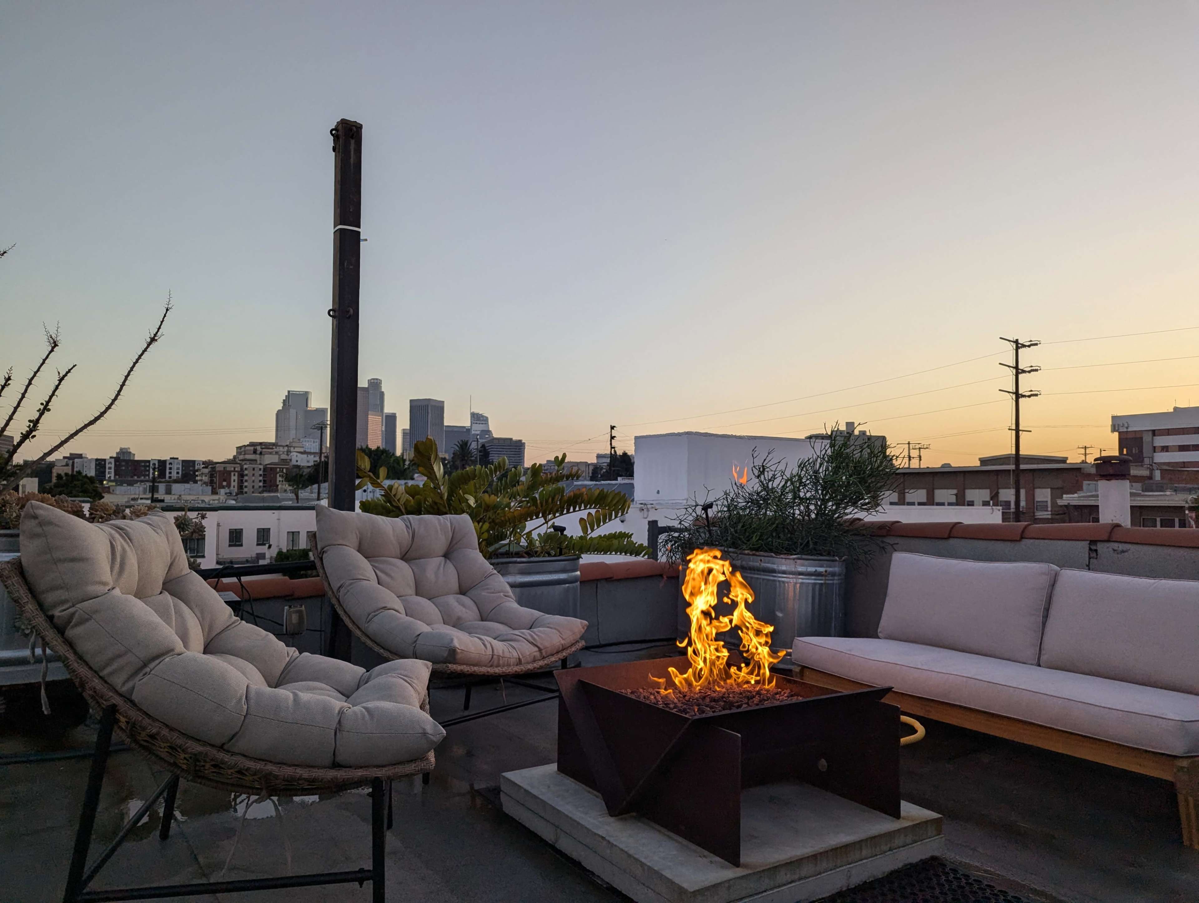 A rooftop seating area with plush chairs and a fire pit overlooks a city skyline at sunset.